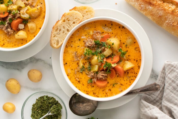 A bowl of creamy soup with potatoes, carrots, celery, ground meat, and herbs, served with a slice of bread. Another bowl, fresh herbs, and a baguette are also on the table. A spoon rests on the bowl’s edge.