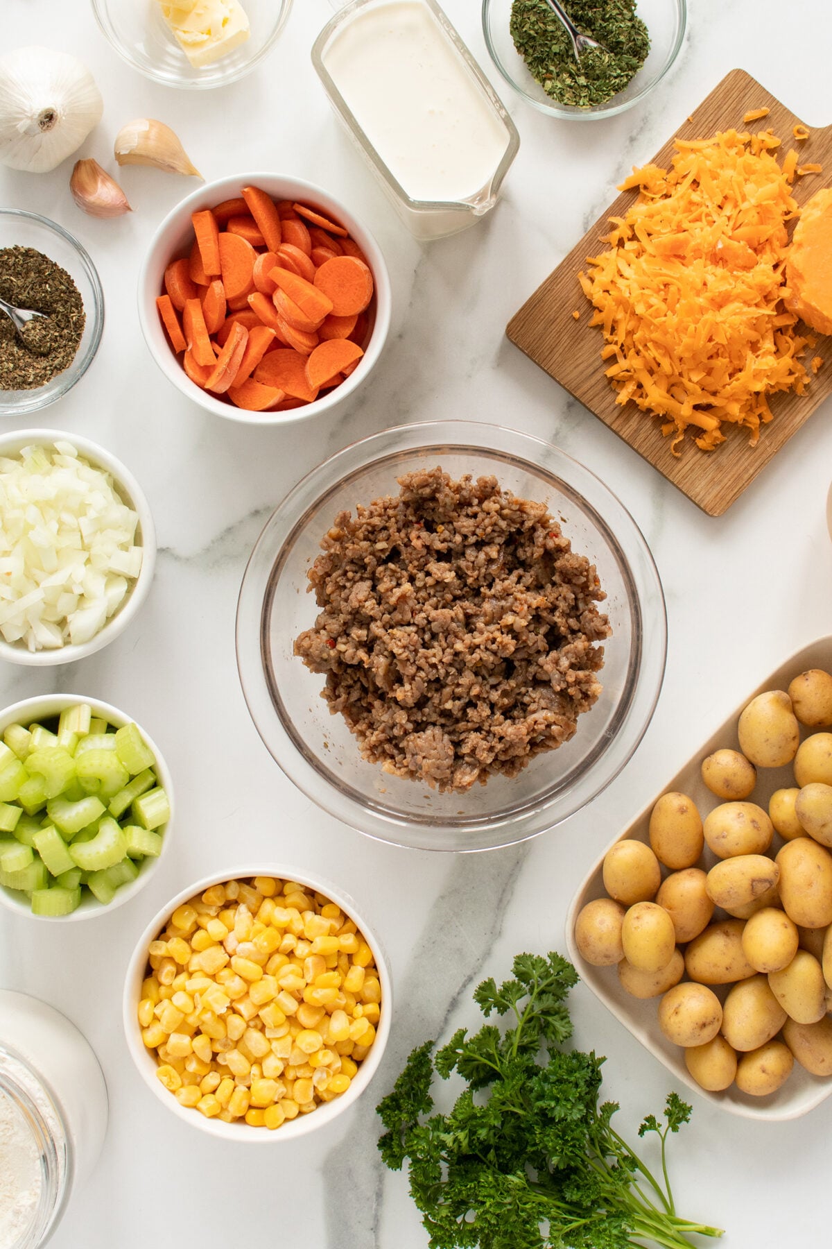 Various ingredients for a recipe are arranged on a white surface, including ground beef, chopped potatoes, grated cheese, carrot slices, corn, celery, onion, parsley, spices, garlic, cream, and butter.