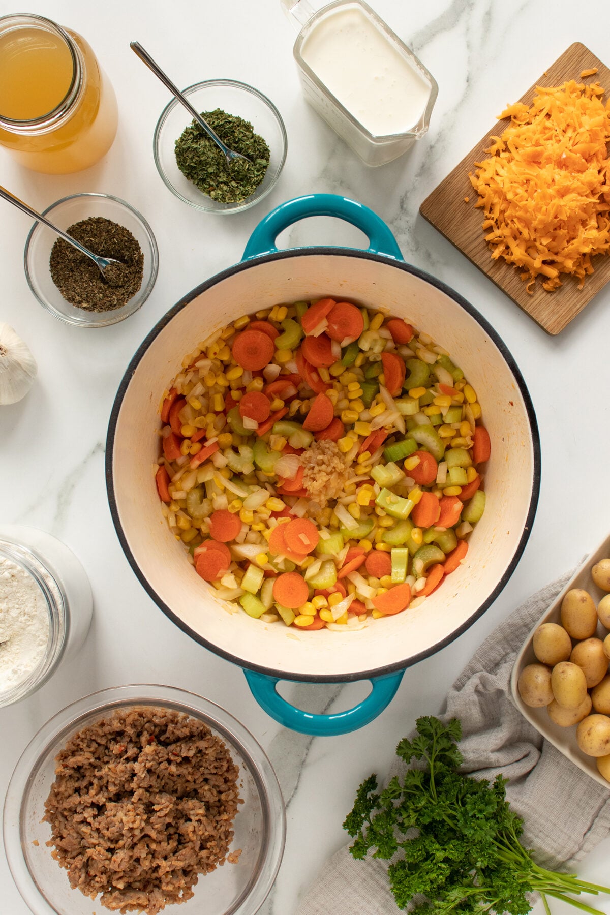 A pot with chopped carrots, celery, onions, corn, and minced garlic sits on a white counter surrounded by small bowls of beef, shredded cheese, seasonings, flour, broth, cream, potatoes, and fresh parsley.