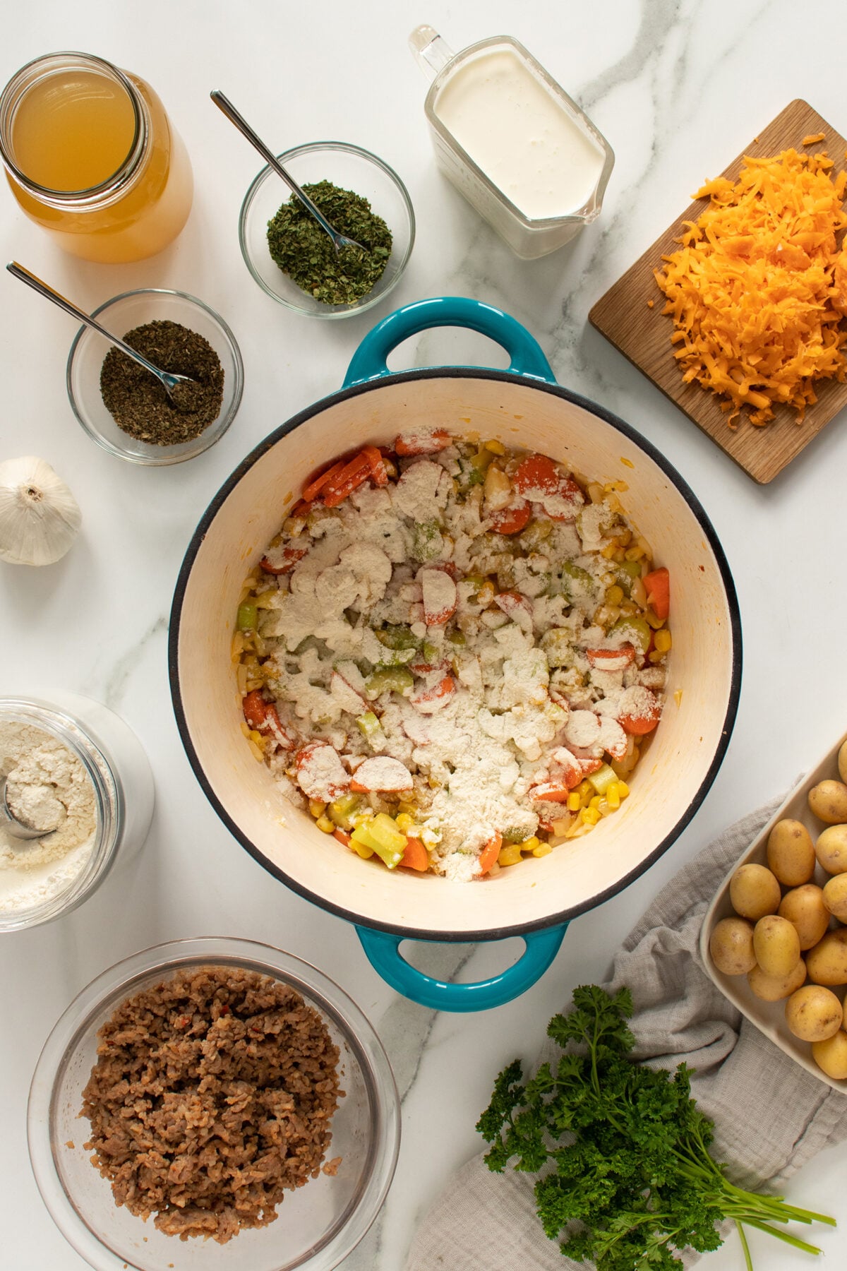 A pot with chopped vegetables and flour sits on a white surface, surrounded by shredded cheese, ground meat, potatoes, fresh herbs, broth, cream, and seasonings, ready for cooking.