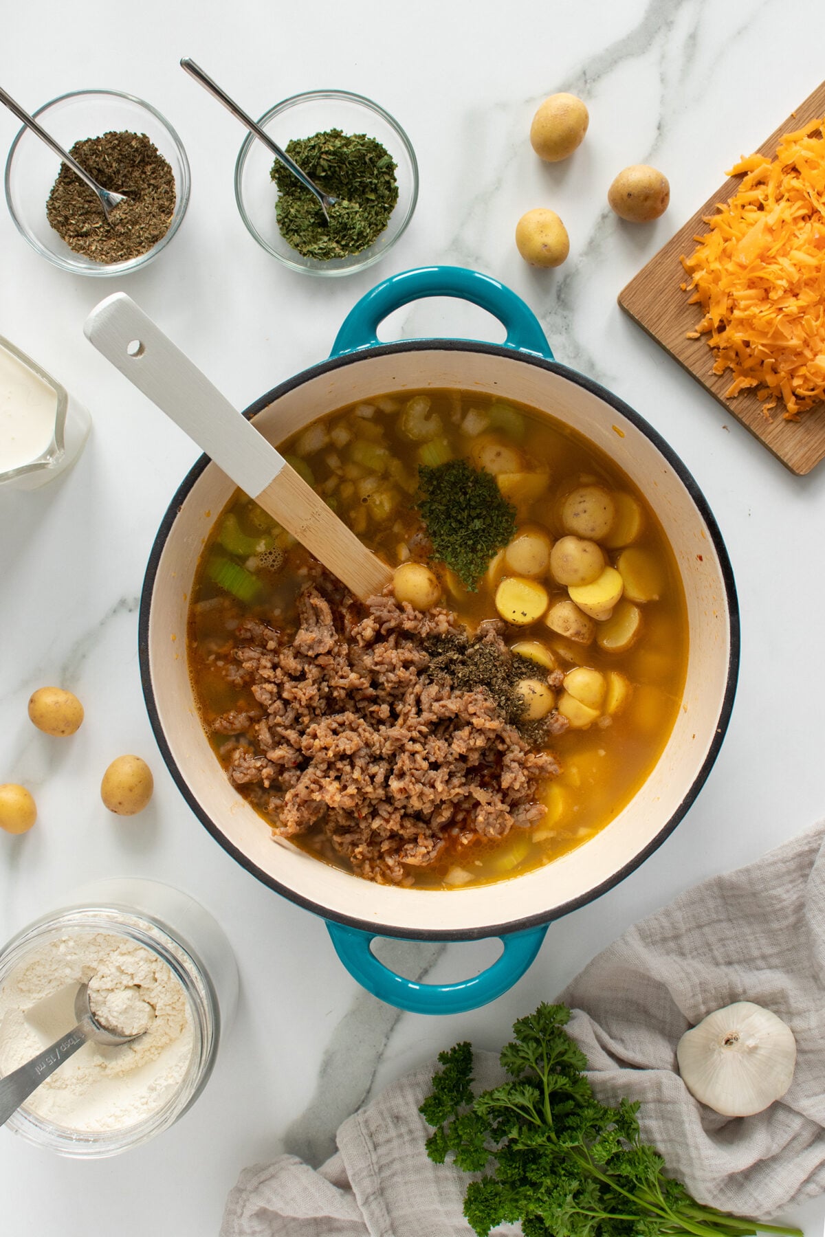 Overhead view of a pot of soup with ground meat, baby potatoes, broth, and herbs, surrounded by shredded cheese, spices, flour, potatoes, parsley, and garlic on a marble countertop.