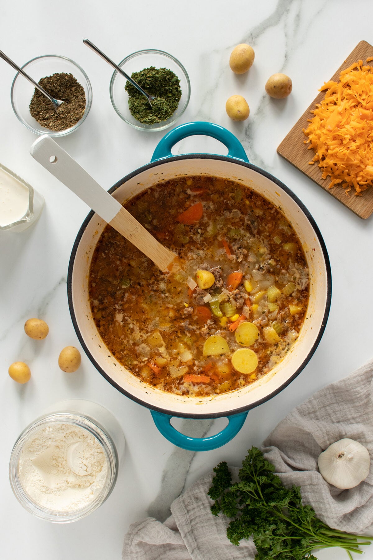 A blue pot filled with soup containing carrots, potatoes, and ground meat sits on a marble counter, surrounded by shredded cheese, fresh herbs, flour, garlic, and small bowls of spices. A white spoon rests in the pot.