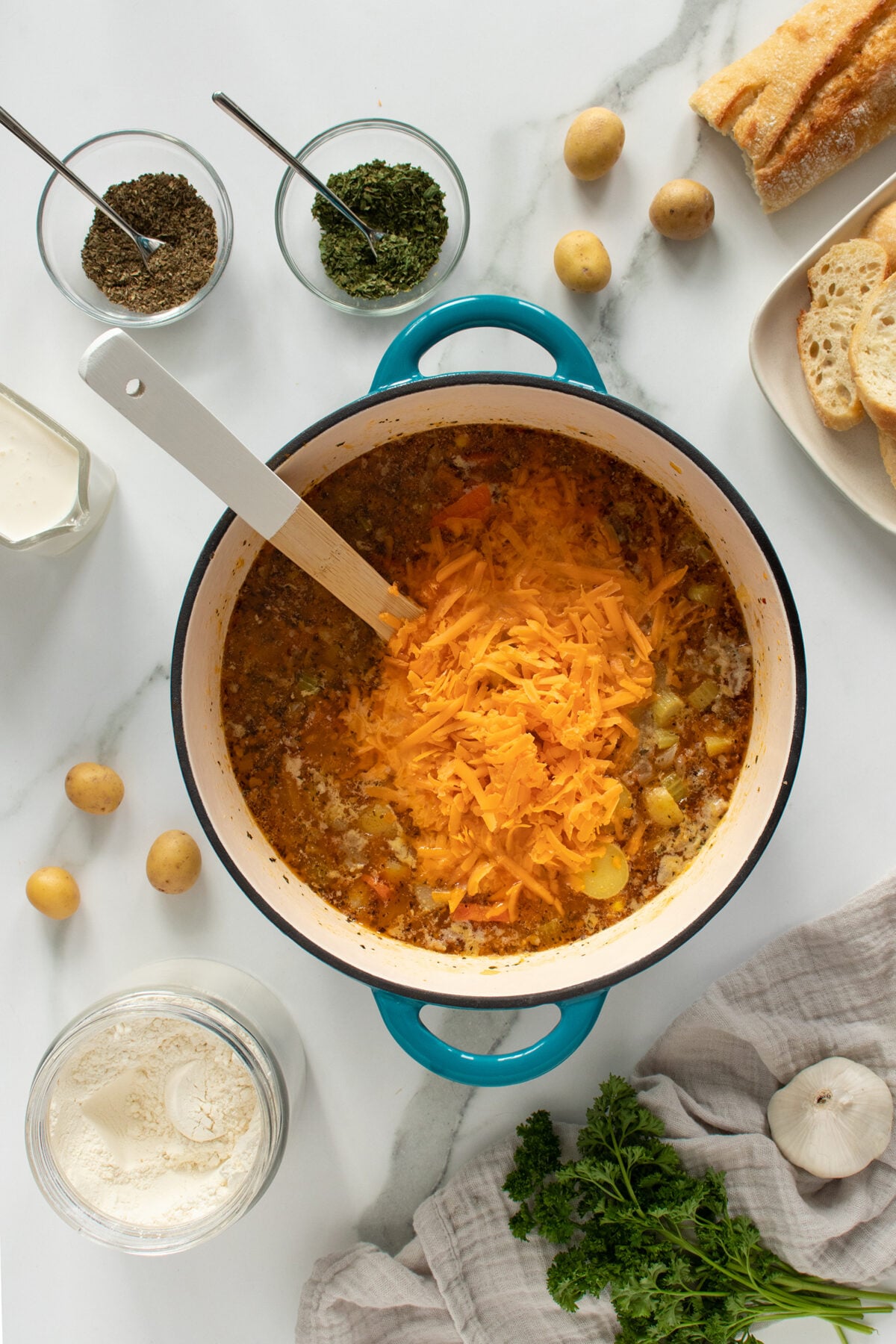 Overhead view of a pot of soup with shredded cheddar cheese on top, surrounded by small potatoes, herbs in bowls, sliced bread, a jar of flour, cream, parsley, and a garlic bulb on a marble surface.