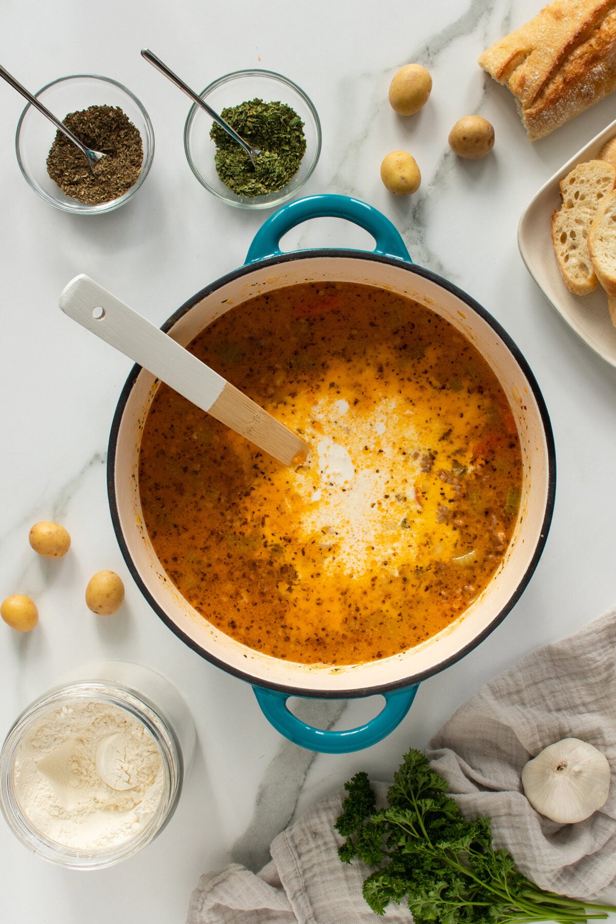 A pot of creamy soup with herbs sits on a white surface, surrounded by potatoes, bread slices, two bowls of dried herbs, fresh parsley, garlic, and a flour jar. A white spoon rests in the pot.