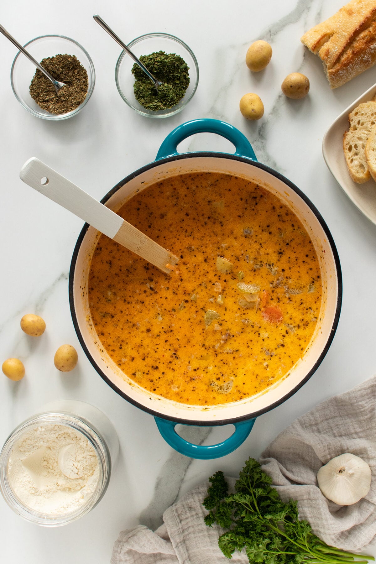 Overhead view of a pot of creamy orange soup with herbs, placed on a white surface surrounded by fresh herbs, garlic, potatoes, sliced bread, flour, and bowls of dried seasonings. A spatula rests in the pot.
