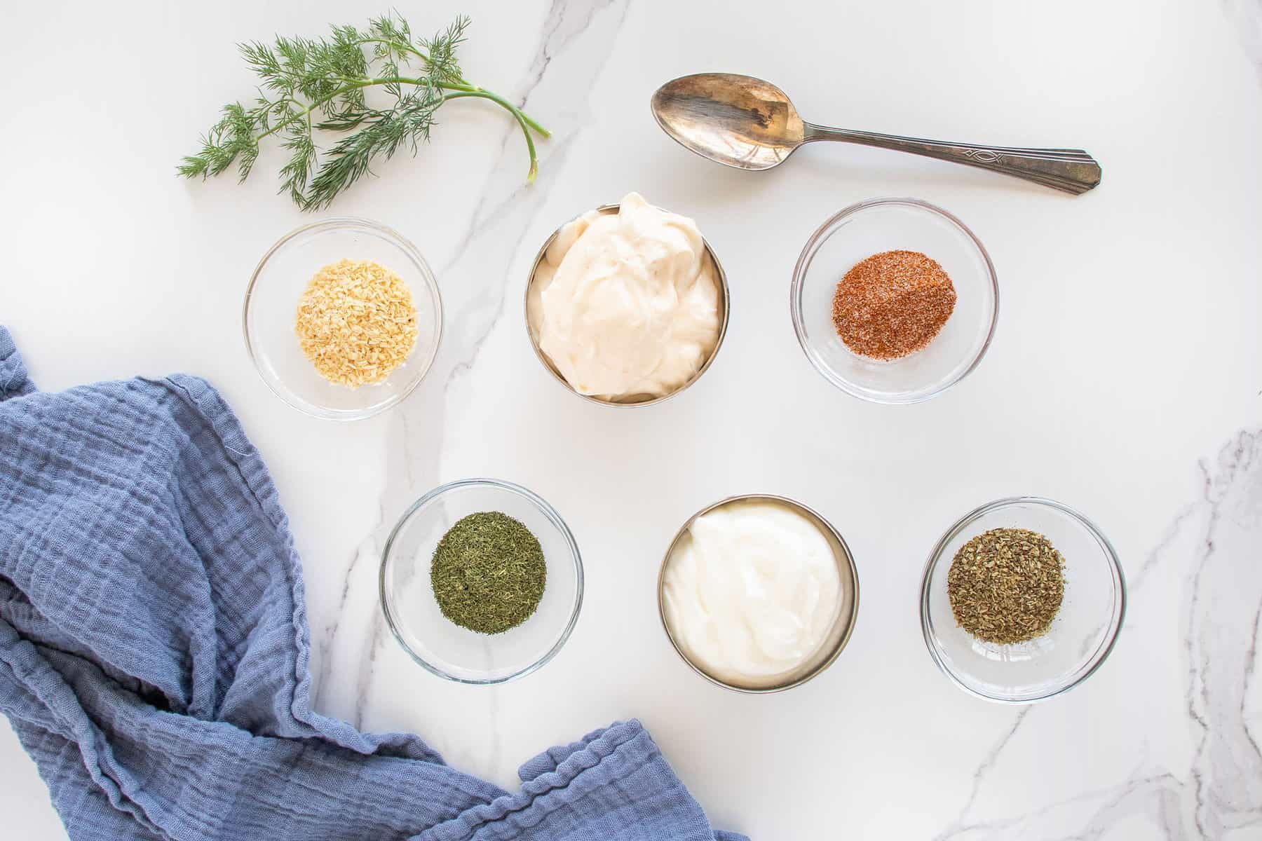 Six small bowls with spices, mayonnaise, and sour cream are arranged on a white marble surface, with a spoon, a fresh dill sprig, and a folded blue cloth nearby.