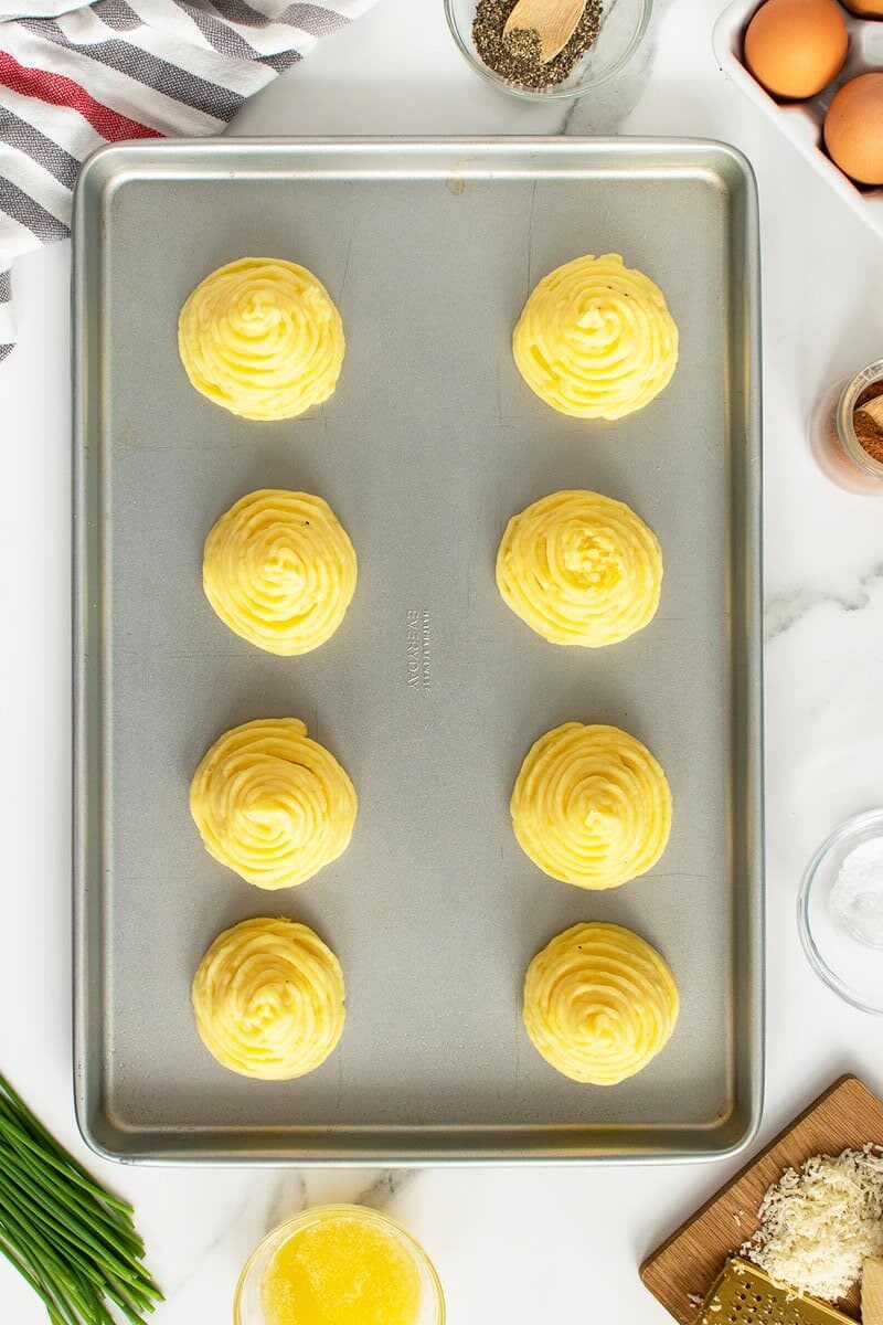A baking sheet with eight piped swirls of mashed potatoes, surrounded by ingredients like eggs, chives, melted butter, cheese, spices, and a striped towel on a white surface.