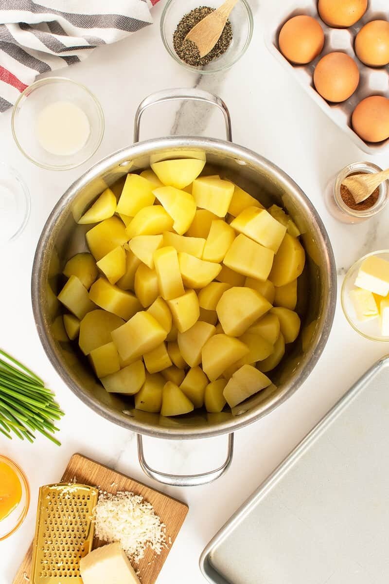 A pot filled with peeled and chopped potatoes is surrounded by eggs, a small bowl of pepper, milk, butter, grated cheese, chopped chives, and a striped kitchen towel on a white countertop.