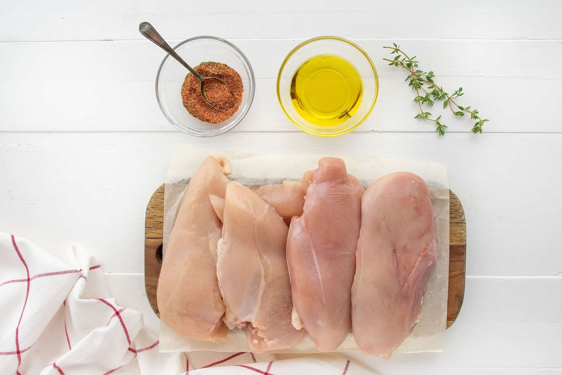 Four raw chicken breasts on parchment paper, placed on a wooden board. Nearby are a bowl of oil, a bowl of spice mix with a spoon, a sprig of fresh thyme, and a red-and-white striped dish towel.
