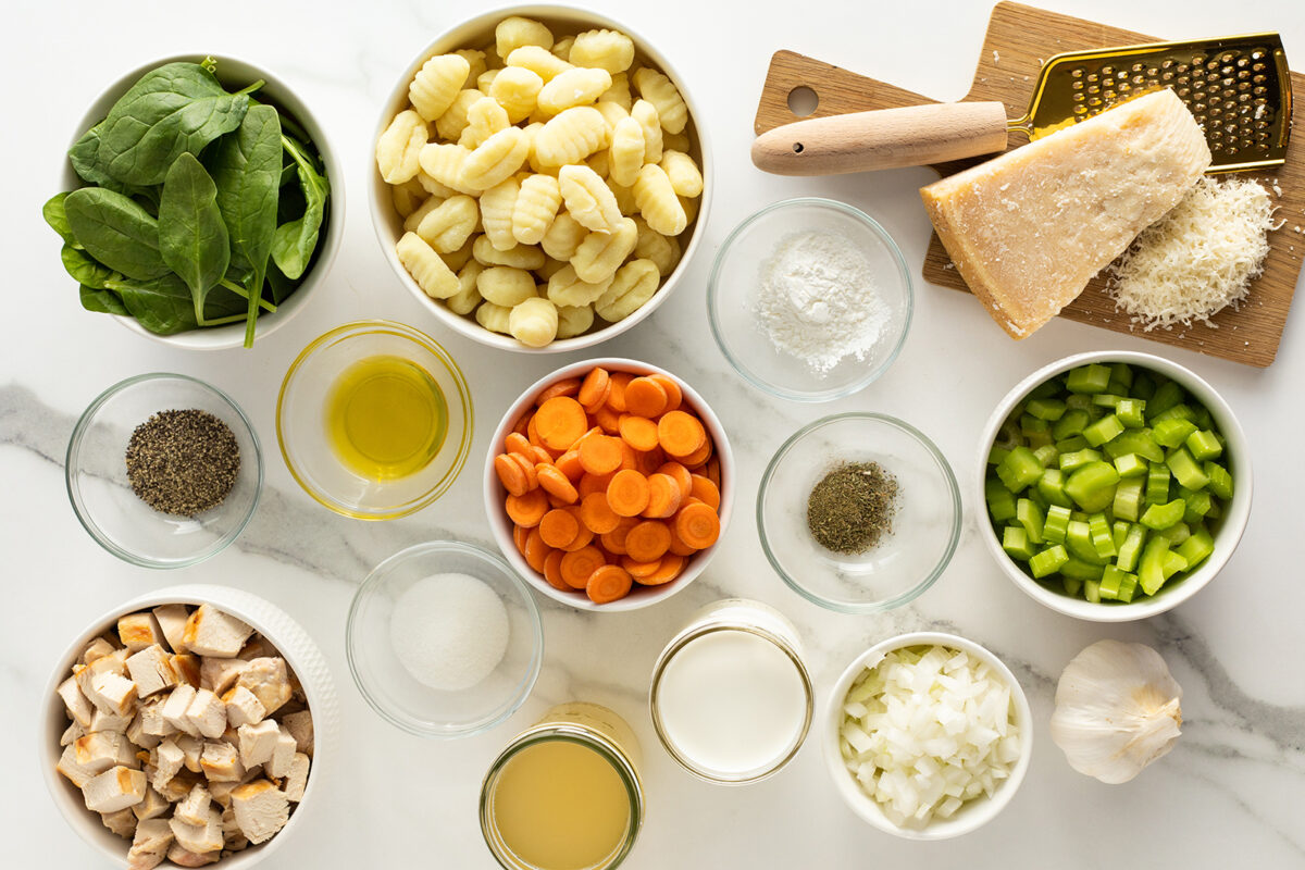 Various ingredients are arranged on a white surface, including bowls of gnocchi, spinach, sliced carrots, chopped celery, cubed chicken, diced onion, seasonings, parmesan cheese, garlic, olive oil, milk, broth, and flour.