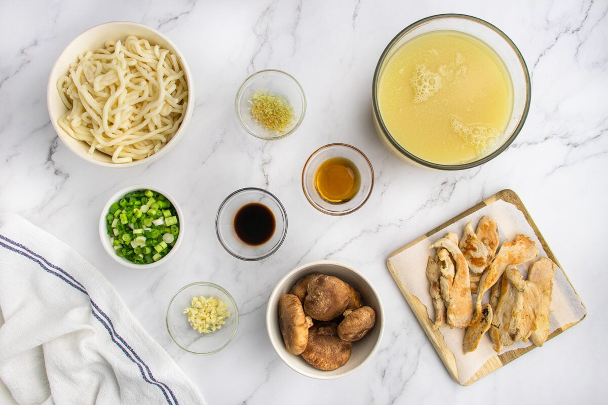 Top-down view of ingredients for a soup: cooked chicken pieces, sliced green onions, shiitake mushrooms, minced garlic, grated ginger, soy sauce, oil, udon noodles, and chicken broth, all arranged on a white marble surface.
