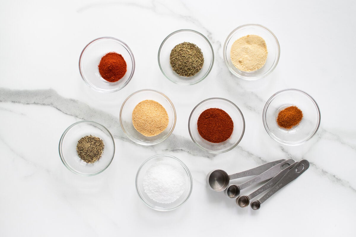 Eight small glass bowls with various ground spices and seasonings are arranged on a white marble surface, along with a set of four metal measuring spoons.