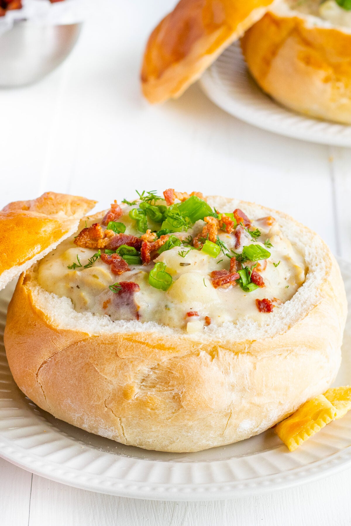 A creamy soup garnished with bacon bits and chopped green onions, served inside a round bread bowl with the top resting to the side, on a white plate.
