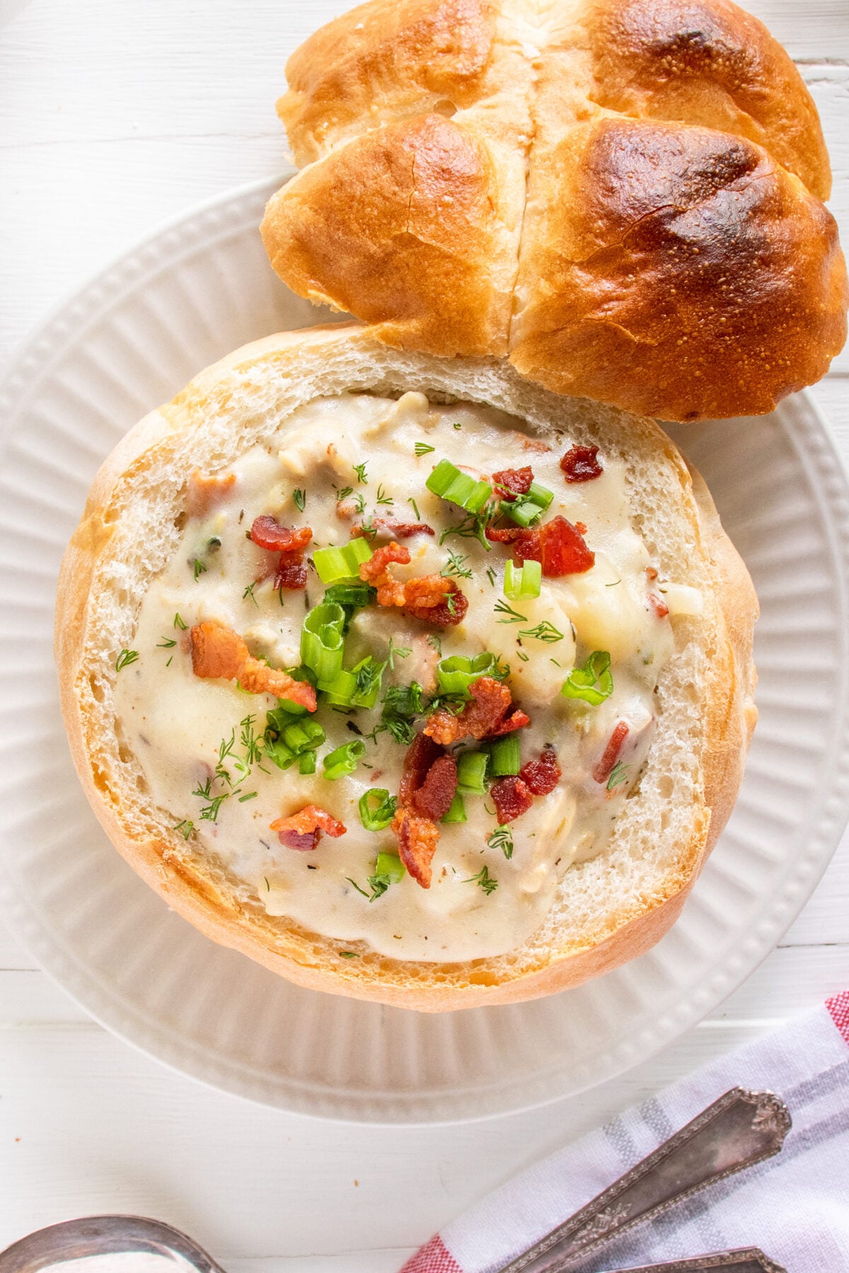 A bread bowl filled with creamy soup, topped with chopped green onions, bacon bits, and herbs, sits on a white plate next to a piece of crusty bread. A spoon and napkin are nearby on a white wooden surface.