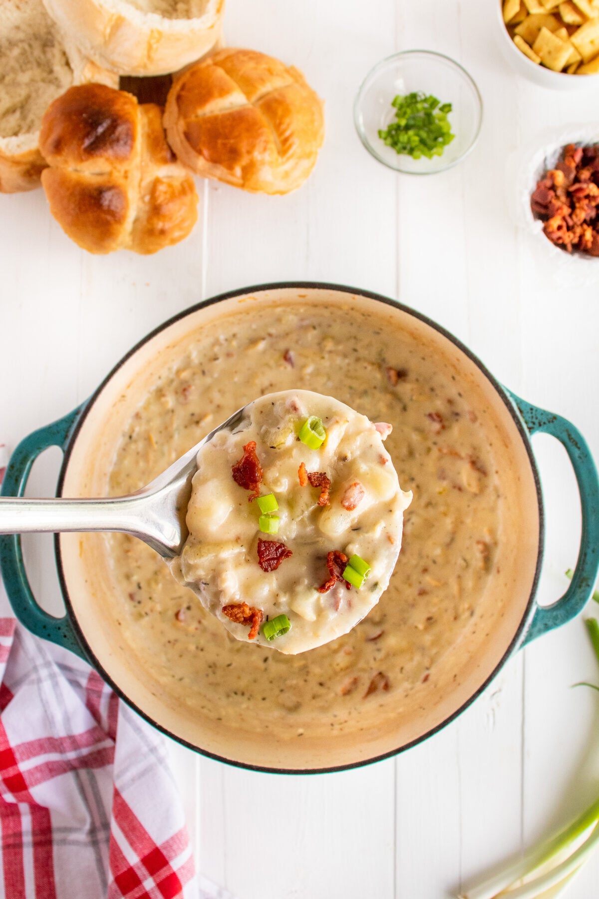 A ladle holds a creamy soup with bacon bits and green onions above a pot. Surrounding the pot are bread rolls, chopped green onions, croutons, and a red-and-white towel on a white table.