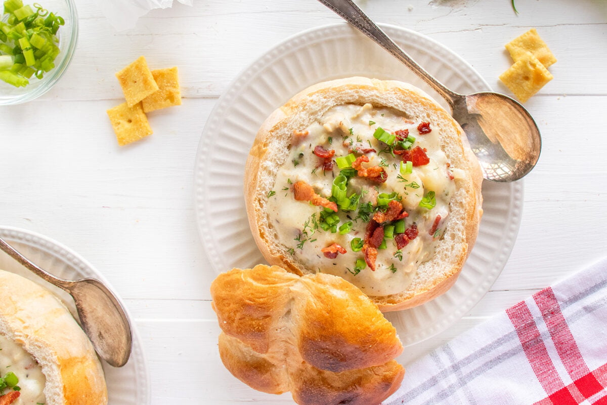 A bread bowl filled with creamy soup topped with bacon bits and chopped herbs, served on a white plate with a spoon and a roll. Crackers and sliced green onions are nearby on a white wooden table.