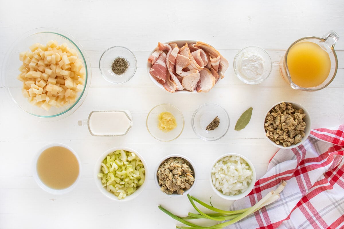 An overhead view of various ingredients arranged neatly on a white surface, including chopped potatoes, bacon, celery, onions, clams, broth, cream, seasonings, and a red checkered kitchen towel.