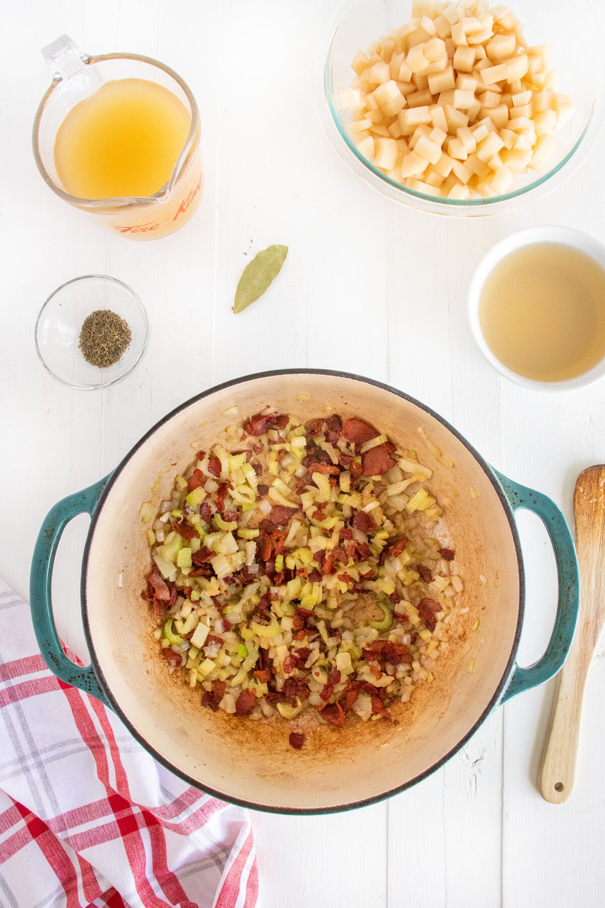 A pot with sautéed onions, celery, and bacon sits on a white surface. Surrounding it are a bowl of diced potatoes, a cup of broth, a bowl of spices, a bay leaf, and a dish of olive oil. A red plaid towel is nearby.