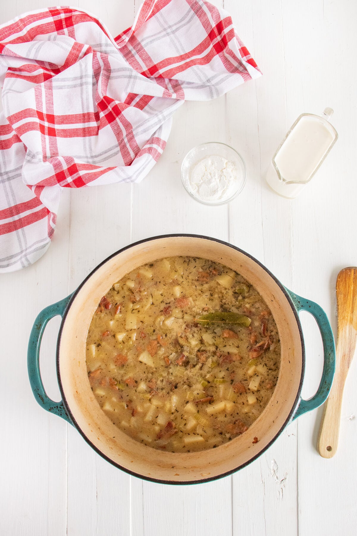 A pot of hearty potato soup with herbs and vegetables sits on a white wooden table. Nearby are a wooden spoon, a red-and-white checkered cloth, a small bowl of flour, and a glass of milk.