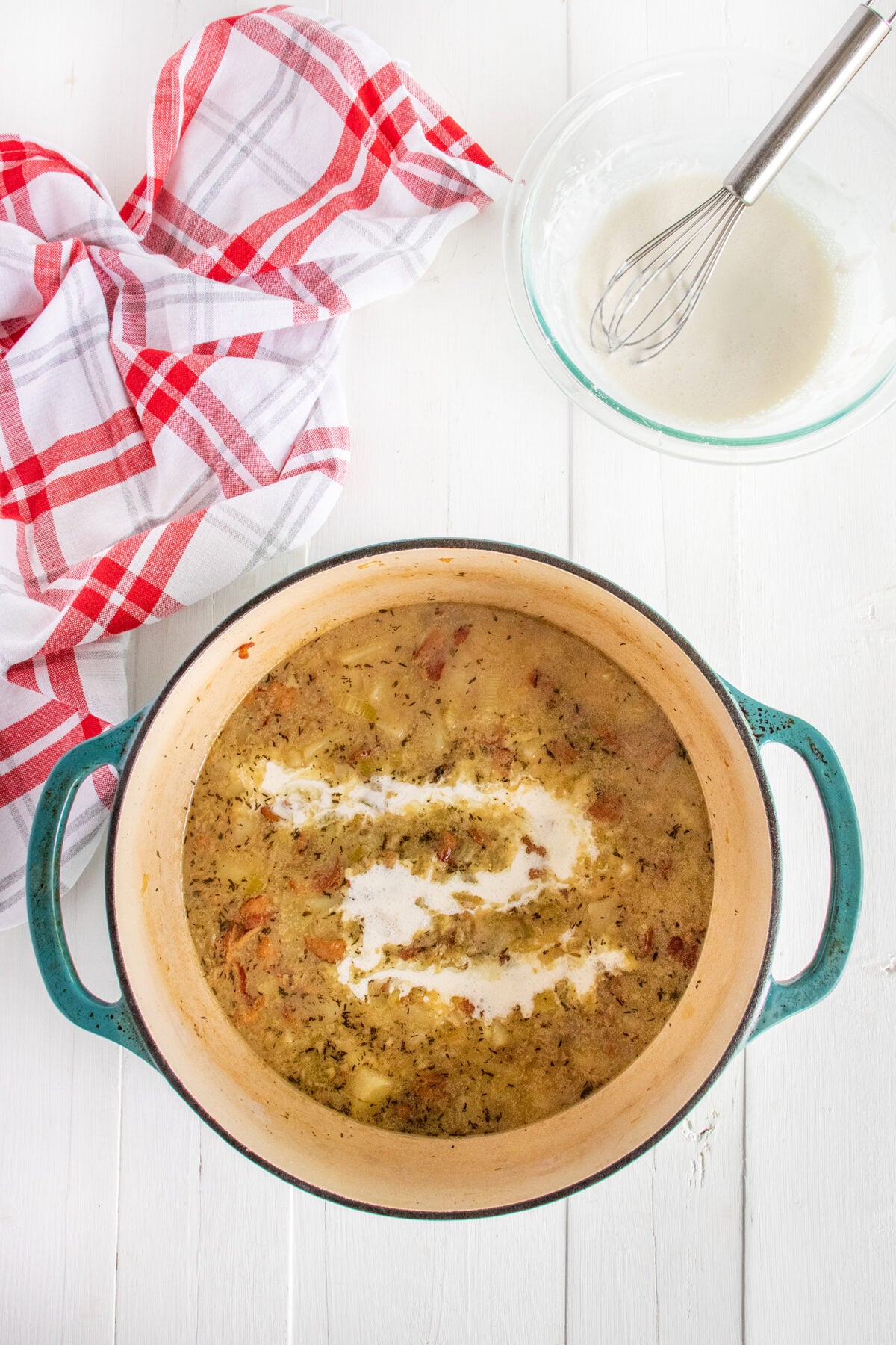 A blue pot filled with creamy soup sits on a white surface, next to a glass bowl with a whisk and a red-and-white checkered towel. A swirl of cream has just been added to the top of the soup.