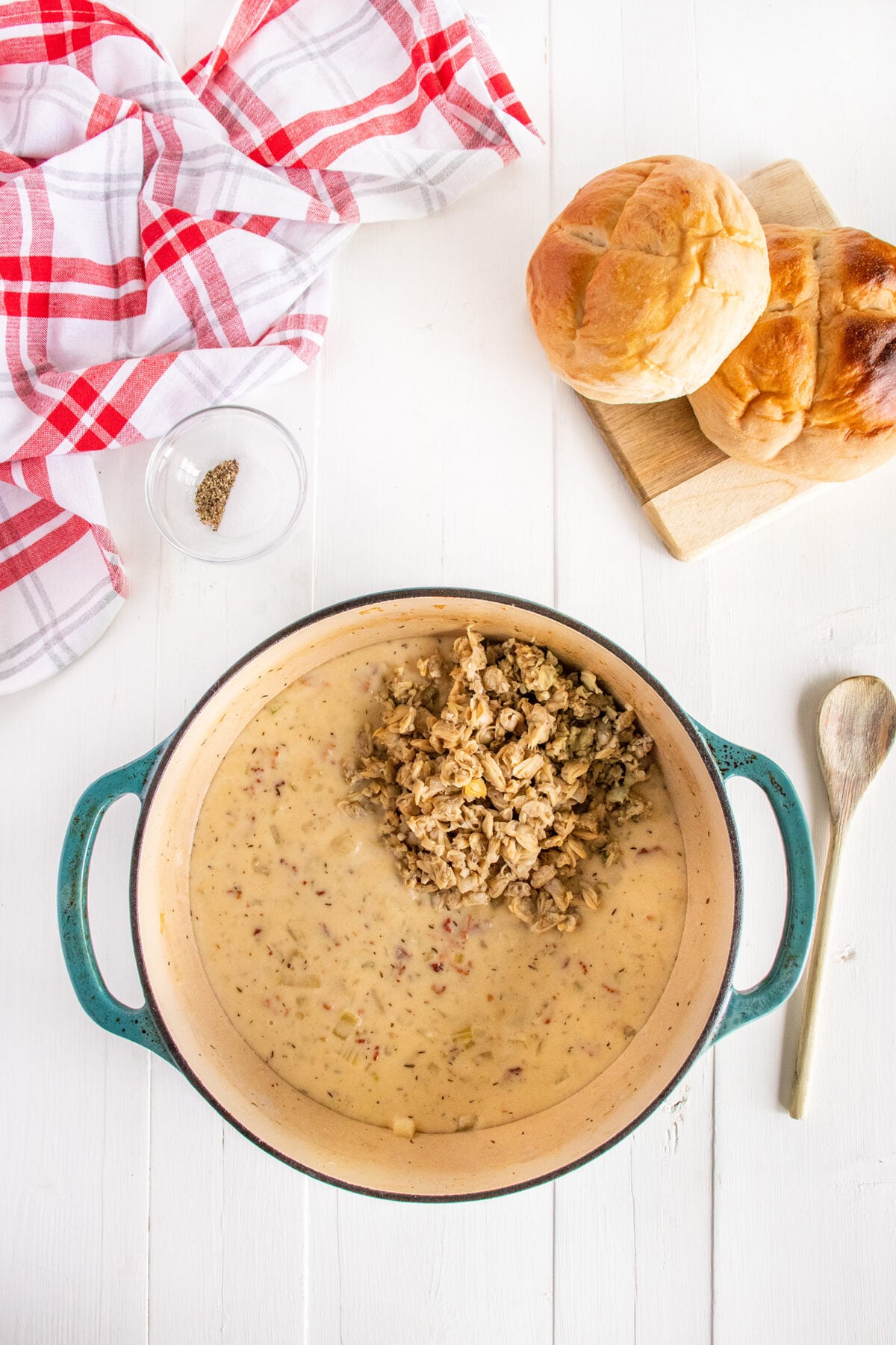 A pot of creamy gravy with crumbled sausage on top sits on a white table beside a wooden spoon, two bread rolls on a cutting board, a small bowl of pepper, and a red-and-white checkered towel.