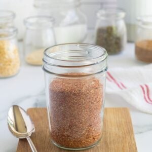 A glass jar filled with reddish-brown spice blend sits on a small wooden board with a spoon next to it. Several jars with various spices and kitchen utensils are in the blurred background.