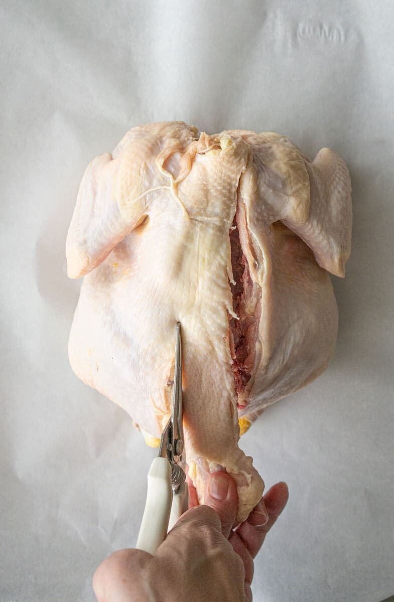 A hand uses kitchen shears to cut along the backbone of a raw whole chicken placed on a sheet of parchment paper.