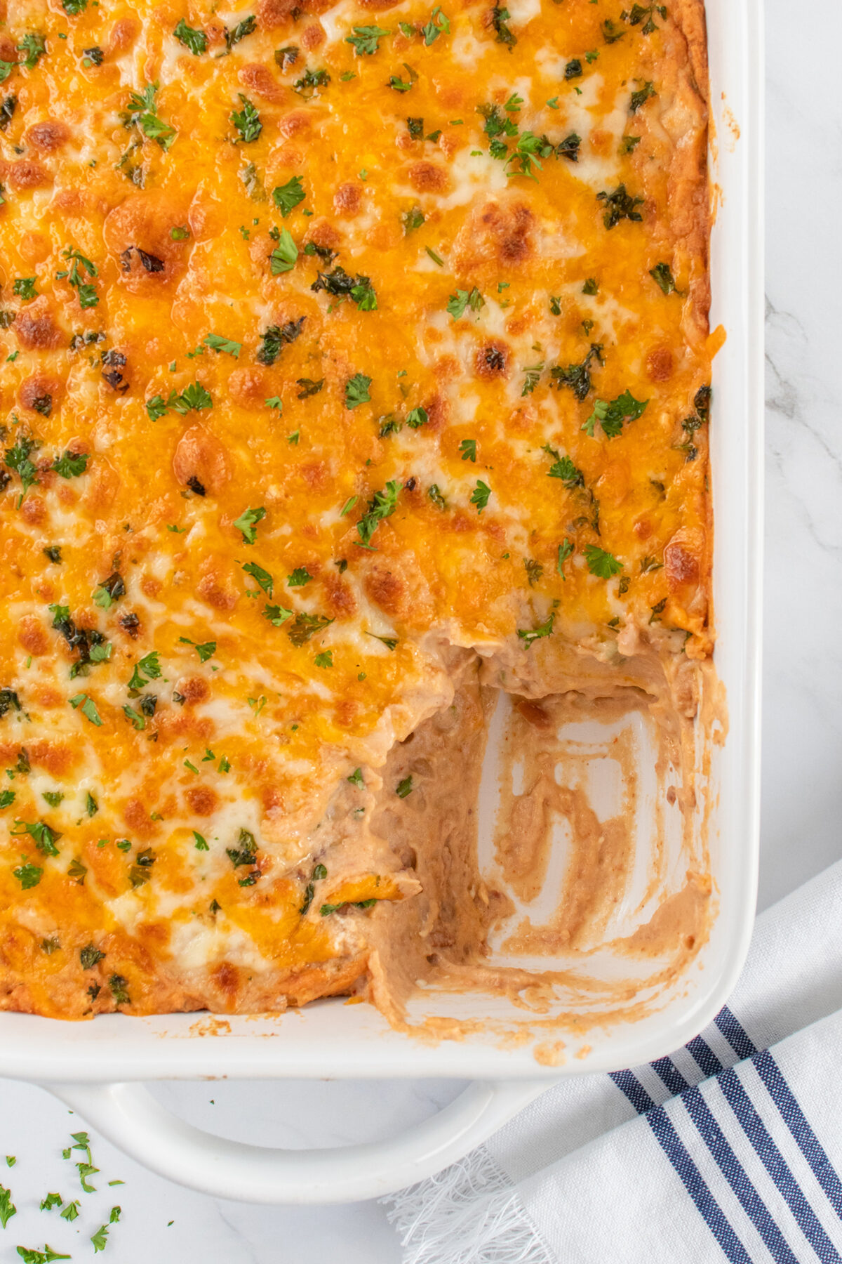 A baked casserole dish with a cheesy, golden-brown top garnished with chopped herbs. A portion has been scooped out, revealing creamy layers underneath. The dish sits on a white marble surface next to a striped towel.