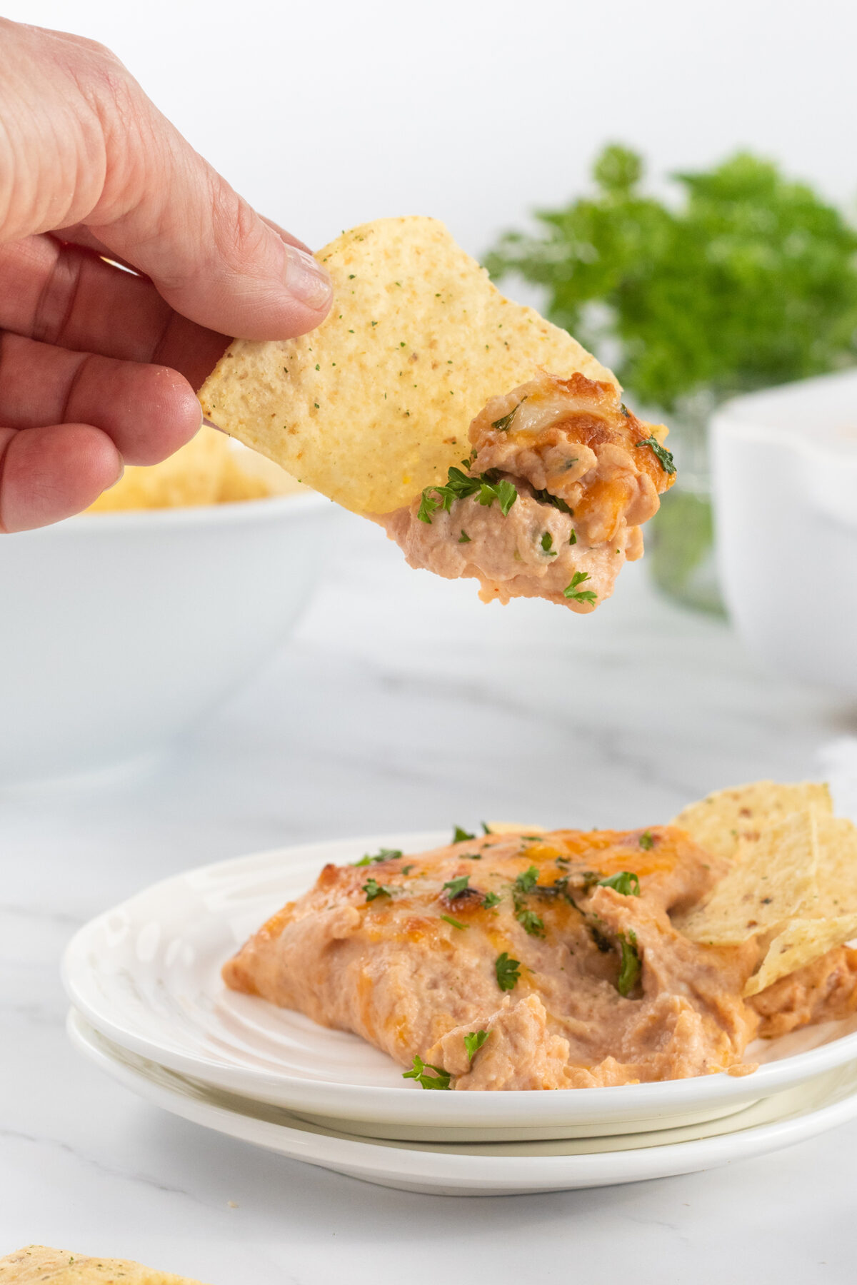 A hand holds a tortilla chip dipped in cheesy bean dip, with more dip and chips on a white plate below. Fresh parsley garnish and a bowl are visible in the background.