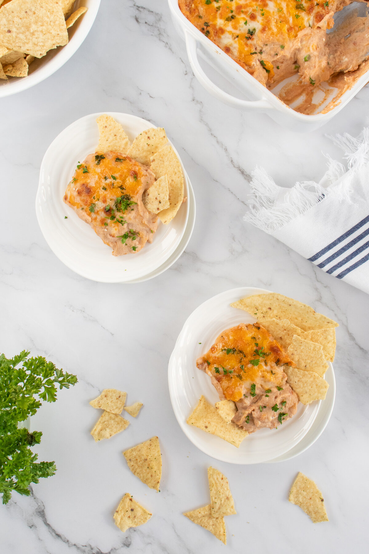 Two small plates with cheesy dip garnished with herbs and served with tortilla chips, placed on a marble surface. A baking dish with more dip and some scattered chips are also visible.