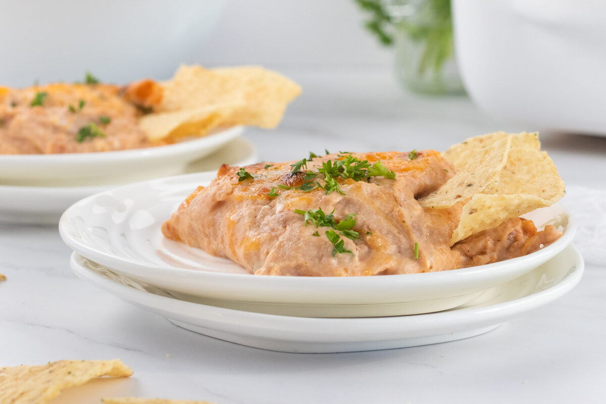 A serving of cheesy bean dip garnished with chopped herbs, accompanied by tortilla chips, on a white plate with another plate of dip in the background.