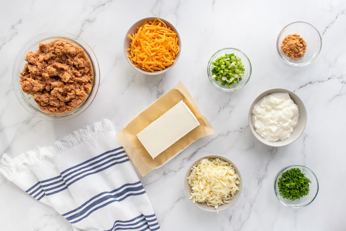 Ingredients on a marble surface include cooked ground meat, shredded cheddar and mozzarella cheese, cream cheese, sour cream, chopped green onions, parsley, and minced garlic, with a striped kitchen towel nearby.
