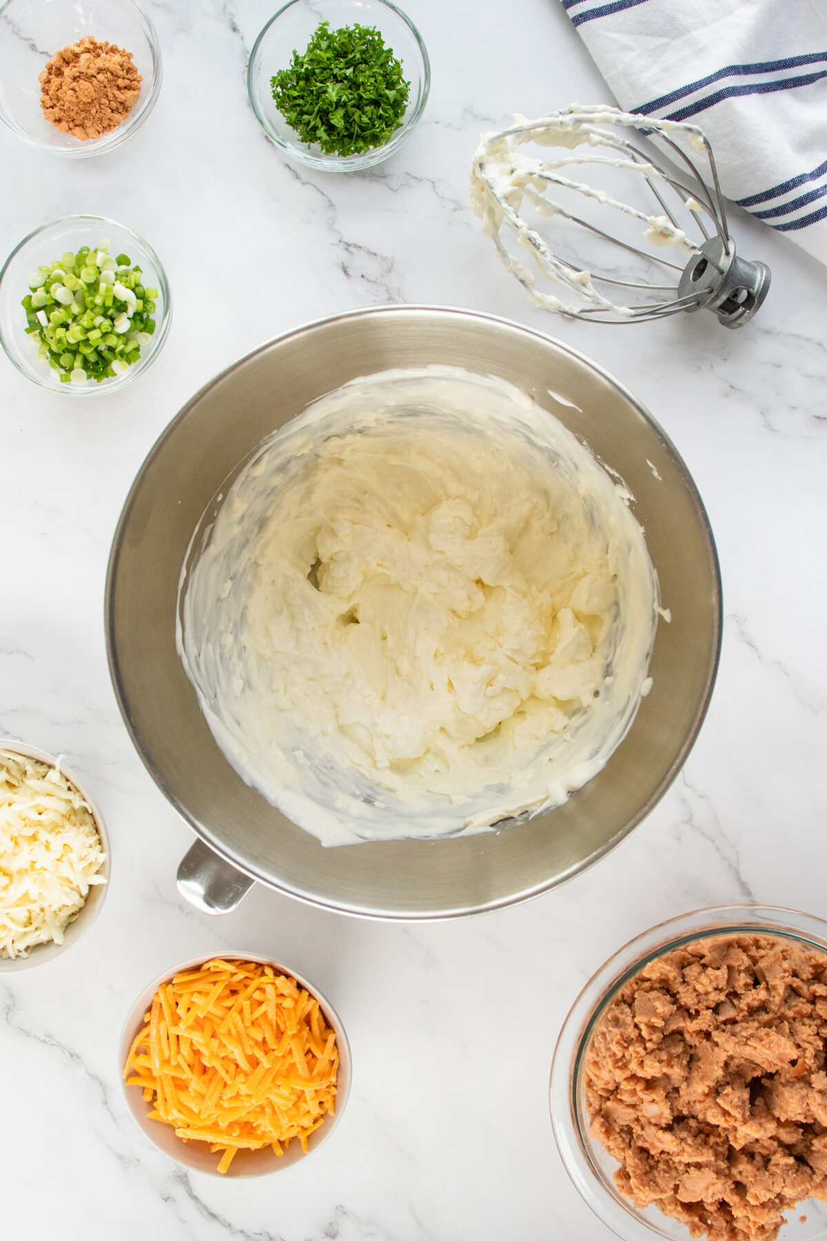 A mixing bowl of creamy mixture surrounded by small bowls containing grated cheese, chopped green onions, parsley, crumbled meat, and a whisk, all on a white marble surface.