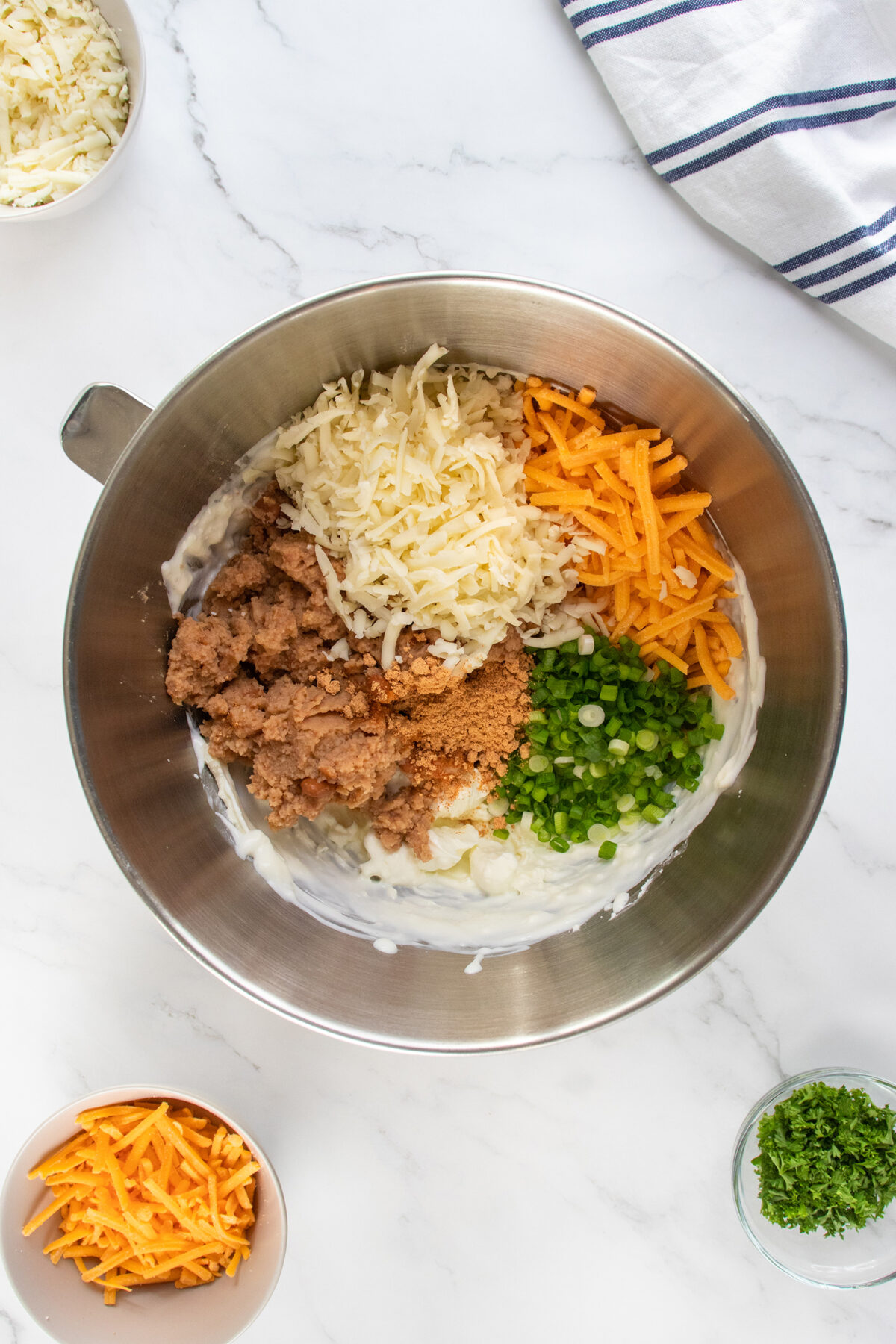 A mixing bowl with shredded cheese, cooked sausage, chopped green onions, and cream cheese, ready to be mixed. Surrounding the bowl are small dishes with more cheese and chopped herbs on a marble countertop.