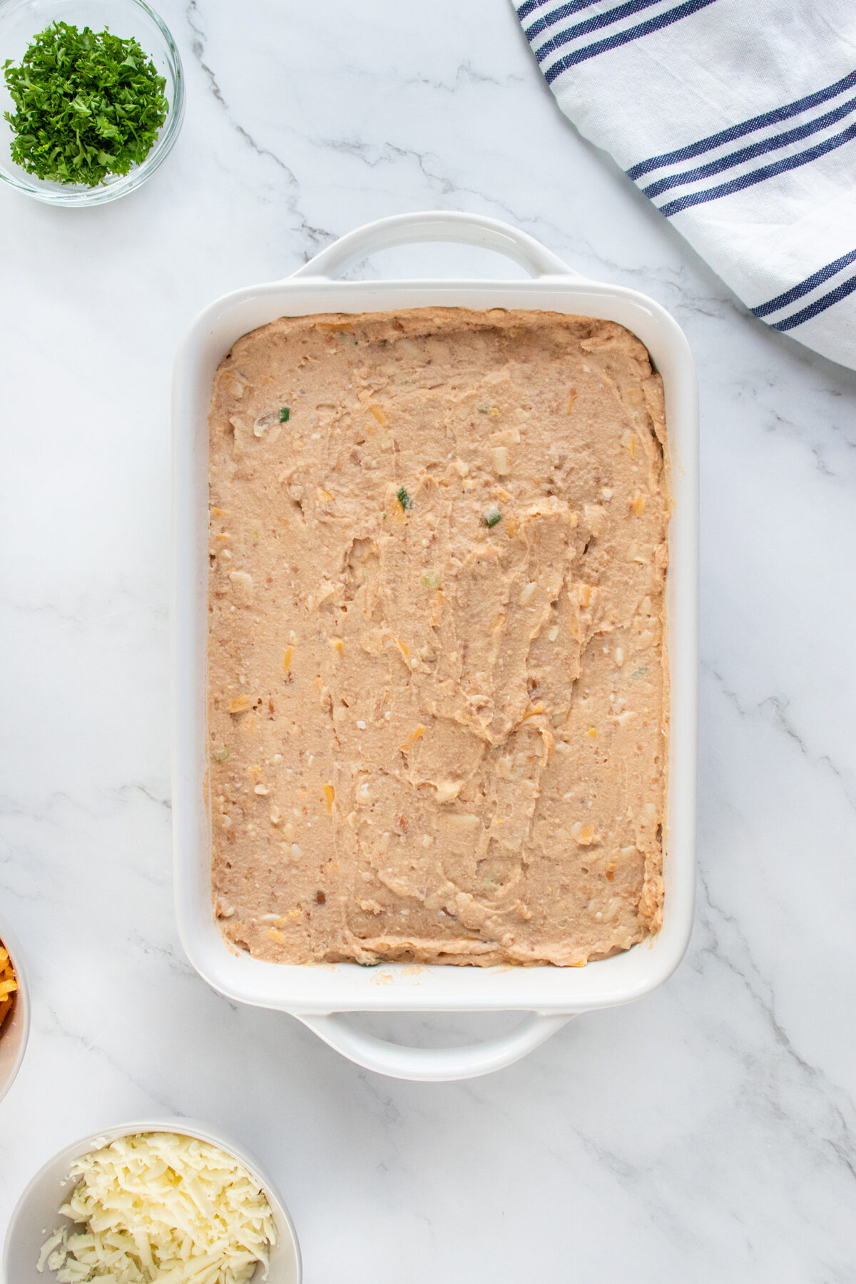A white rectangular baking dish filled with a smooth, spread refried bean mixture sits on a marble countertop. Nearby are bowls of chopped herbs and shredded cheese, and a striped kitchen towel.