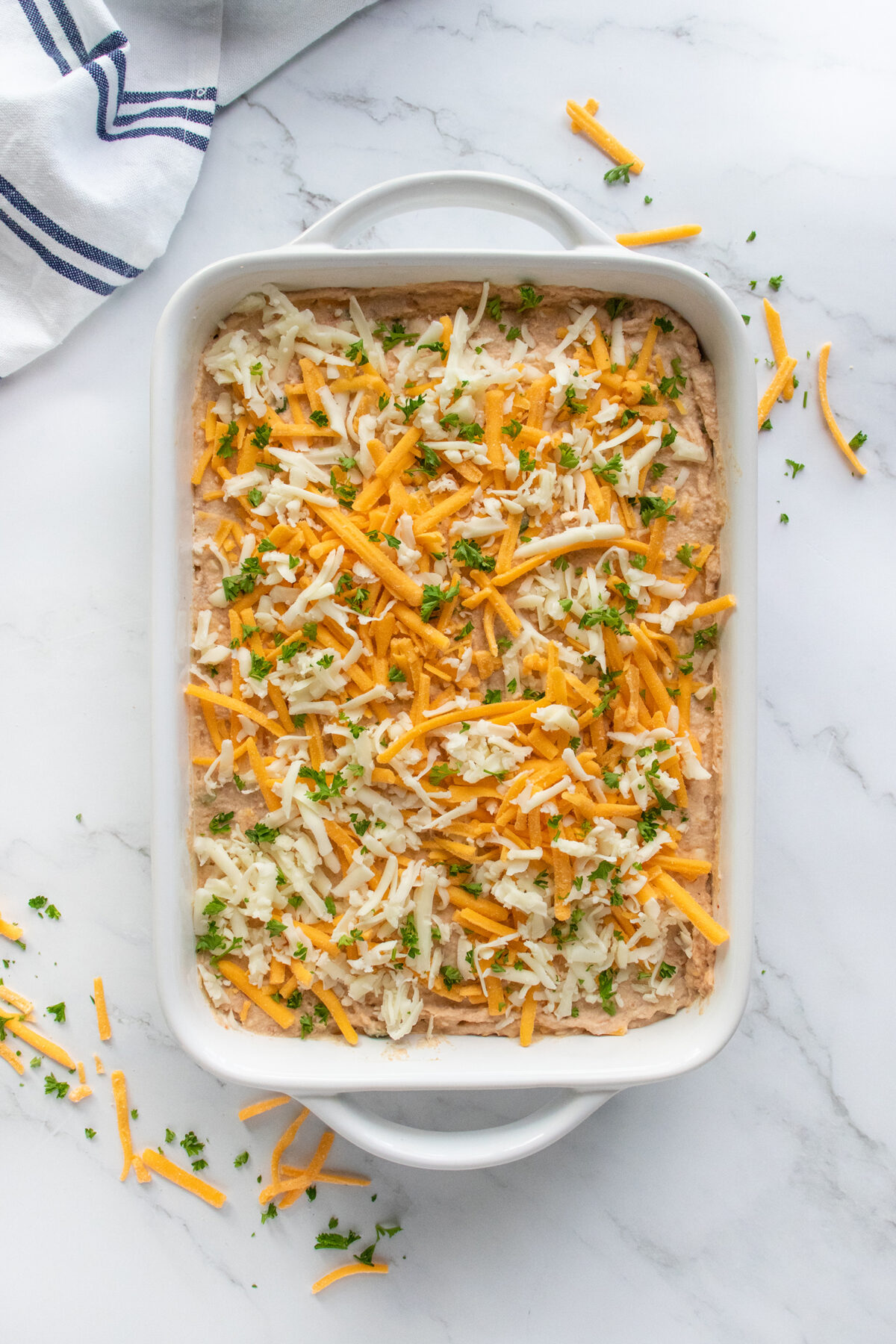 A white baking dish filled with a creamy mixture topped with shredded cheddar and mozzarella cheese, garnished with chopped parsley, on a marble surface with a striped towel nearby.