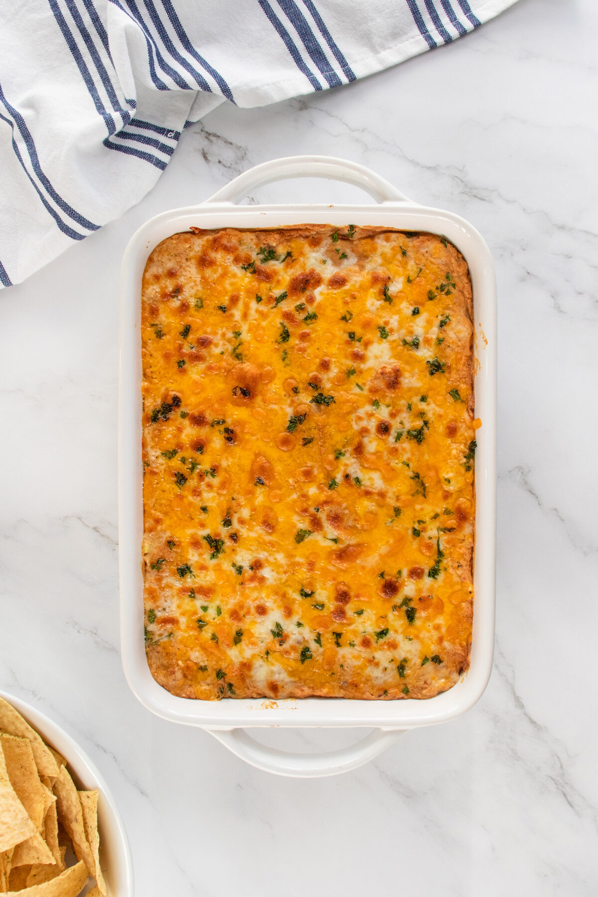 A casserole dish filled with hot, baked cheesy dip topped with melted cheese and herbs, sitting on a marble surface beside a bowl of tortilla chips and a striped kitchen towel.