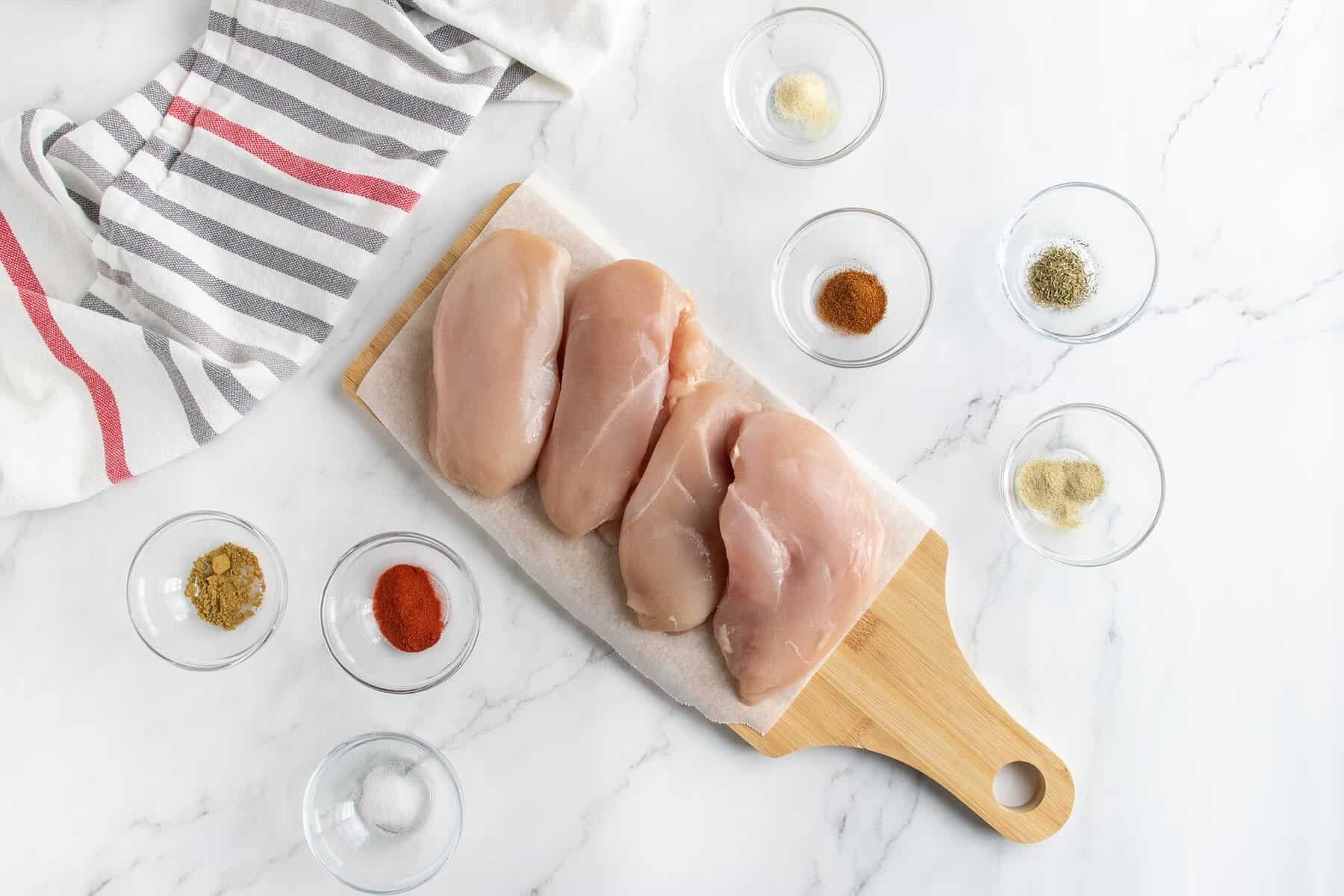 Four raw chicken breasts on a wooden cutting board, surrounded by small glass bowls of various spices and a striped kitchen towel on a white marble surface.
