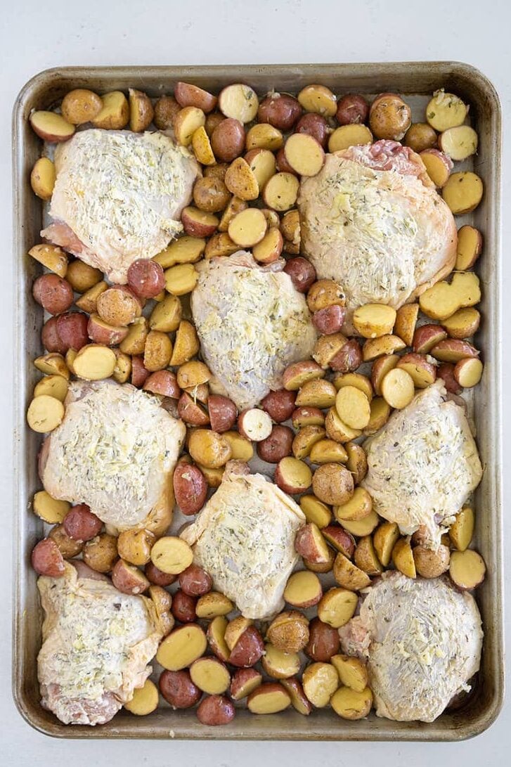 A baking sheet filled with seasoned raw chicken thighs and halved baby potatoes, evenly spread out and ready to be roasted.