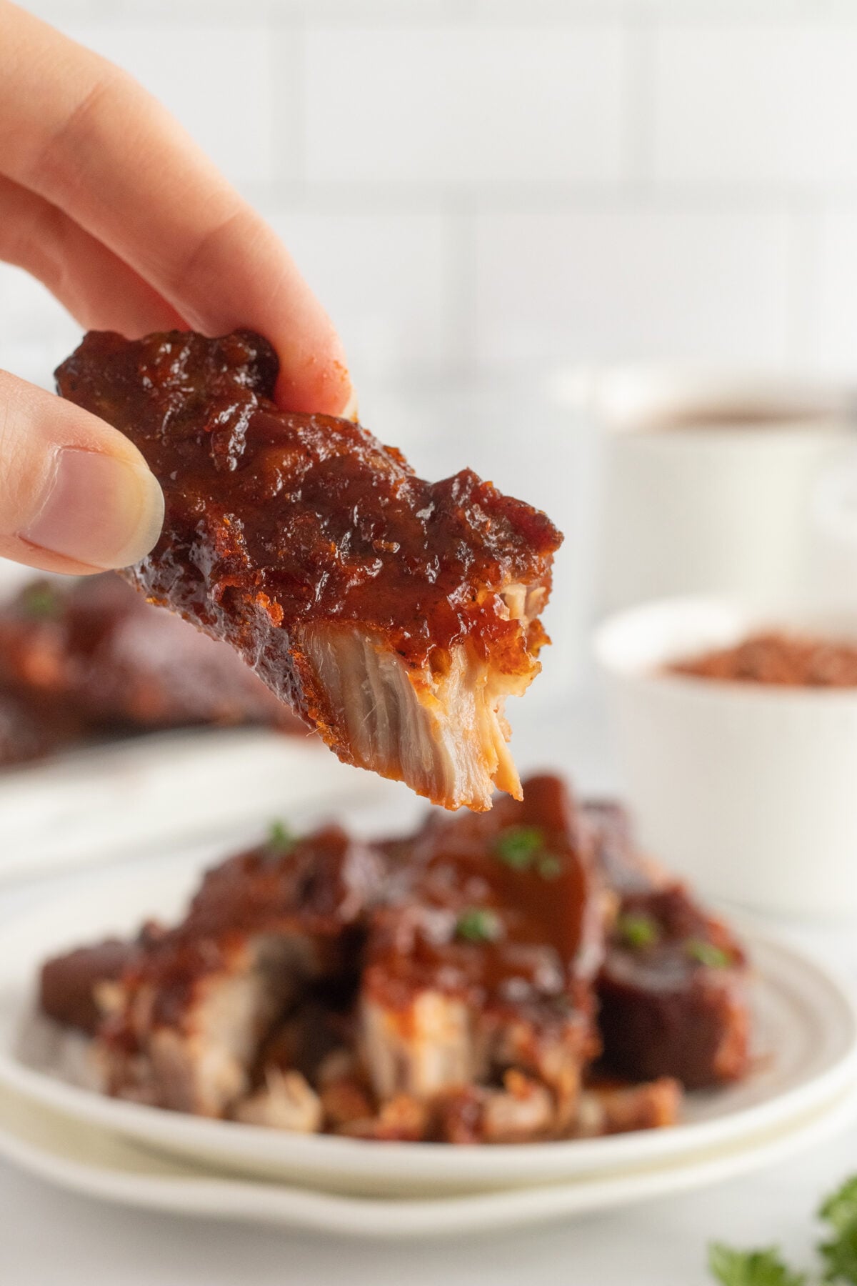 A hand holds up a saucy, tender rib with a bite taken out, while a plate of ribs garnished with herbs sits blurred in the background.