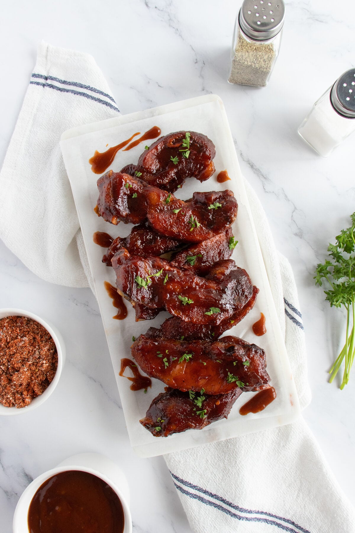 A rectangular white plate of glazed BBQ ribs garnished with chopped parsley, placed on a striped towel. Nearby are small bowls of seasoning and sauce, salt and pepper shakers, and fresh parsley on a marble surface.