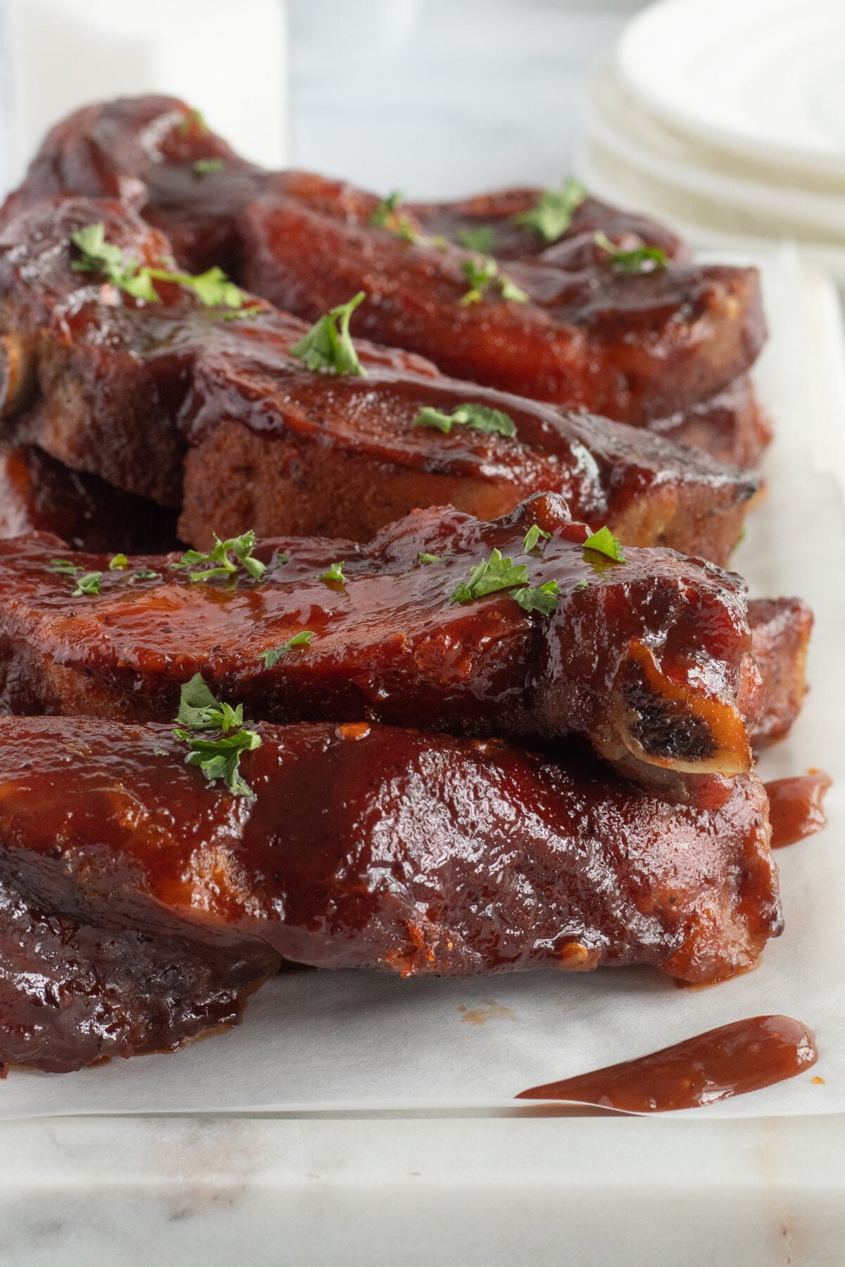 A close-up of barbecue ribs covered in a thick, glossy sauce, garnished with fresh chopped parsley, served on a white surface with stacked plates blurred in the background.