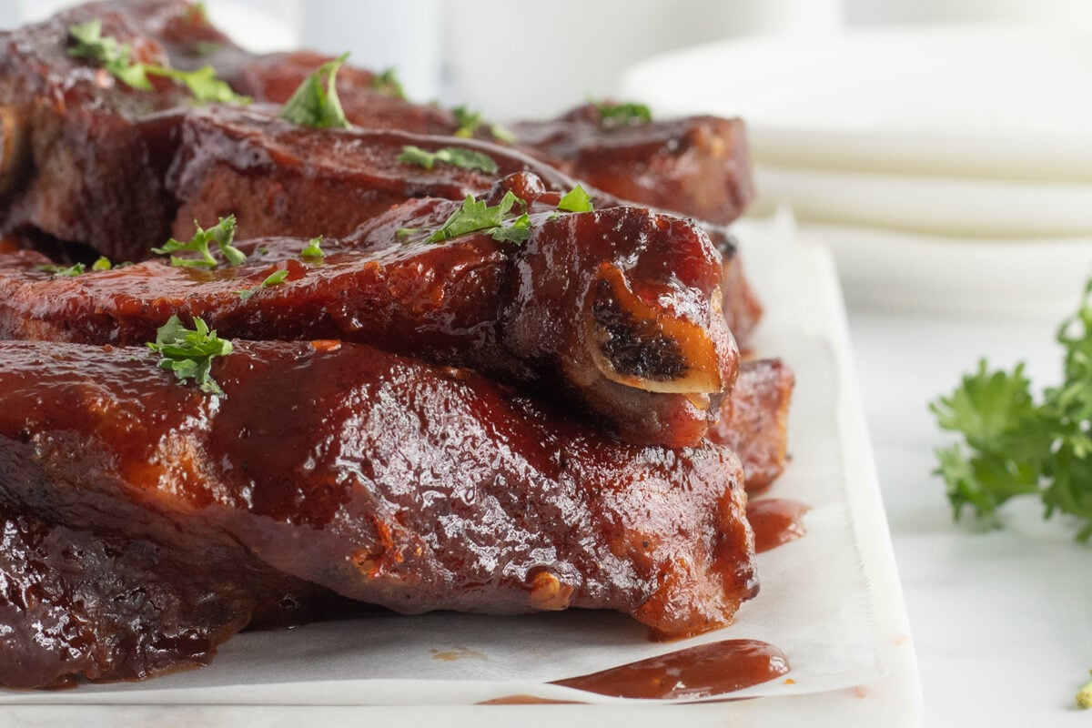 Close-up of juicy, glazed barbecue ribs stacked on a white plate, garnished with fresh chopped parsley. The sauce is thick and shiny, with some dripping onto the plate. White dishes and greenery are blurred in the background.