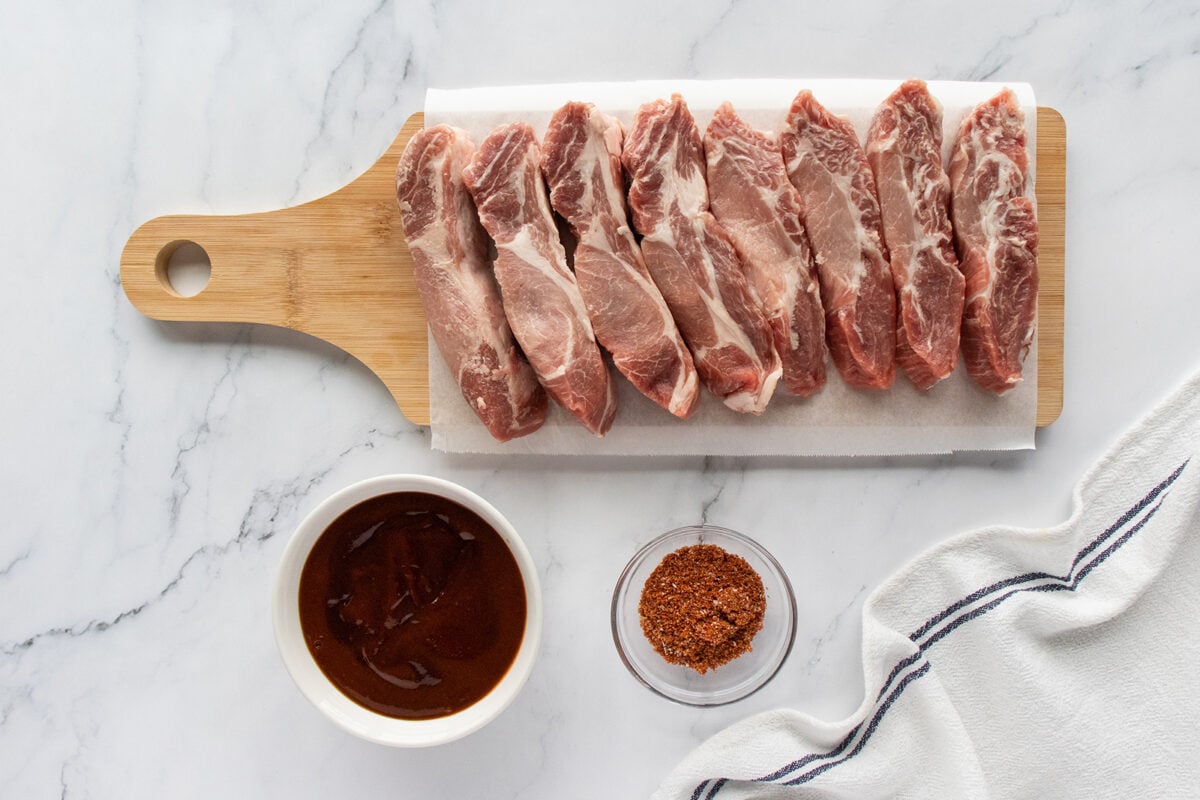 Raw pork ribs are neatly arranged on a wooden cutting board lined with parchment paper. Next to them are a bowl of barbecue sauce, a bowl of dry spice rub, and part of a striped kitchen towel on a marble surface.