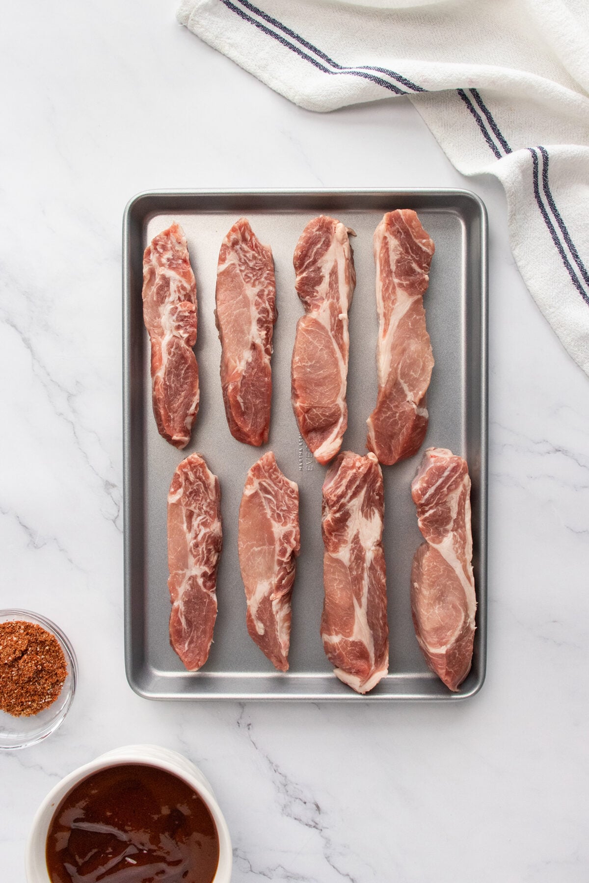 A metal baking sheet with eight raw boneless pork ribs arranged neatly in two rows on a marble surface. Nearby are a bowl of barbecue sauce, a bowl of spice rub, and a white kitchen towel.