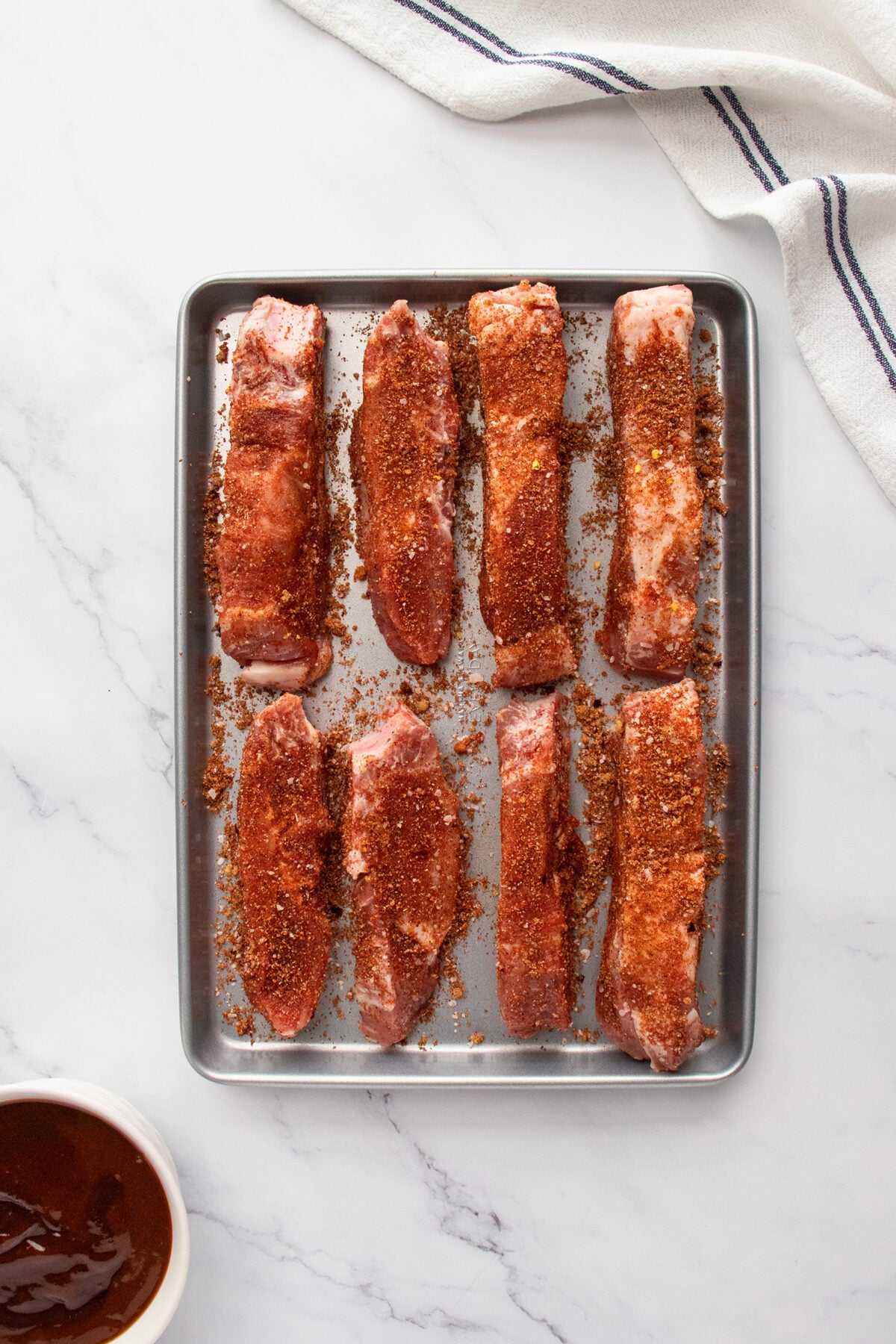 A metal baking tray holds eight raw pork ribs coated with a reddish-brown spice rub. The tray is on a white marble surface next to a bowl of sauce and a white kitchen towel with blue stripes.