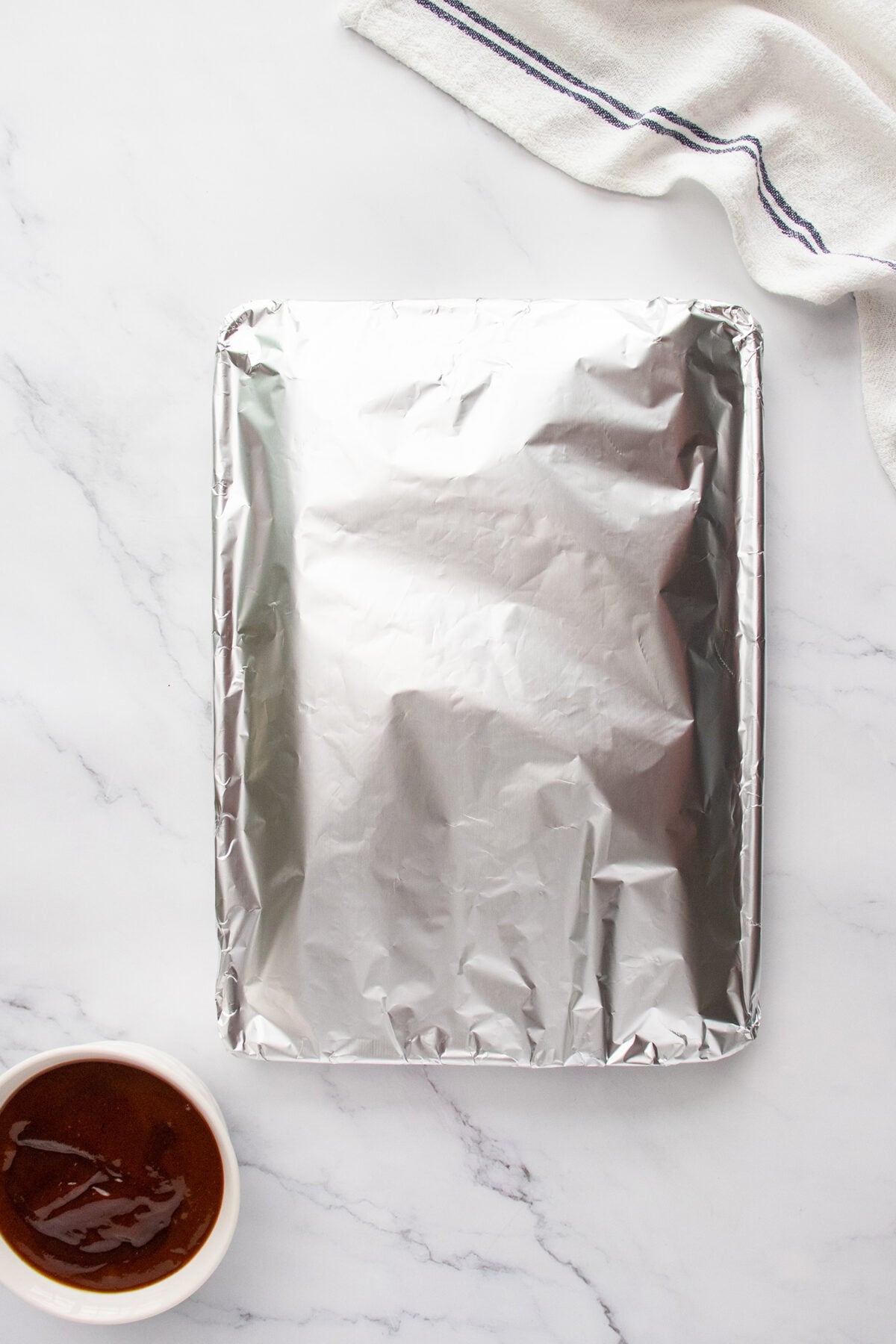 A baking tray covered with aluminum foil sits on a marble surface, with a small bowl of sauce nearby and a folded white kitchen towel in the corner.