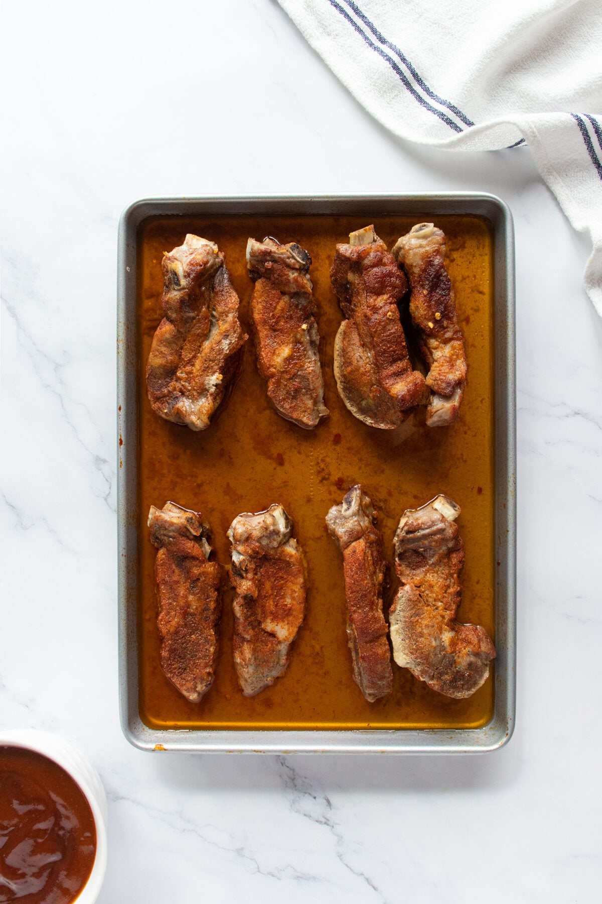 A metal baking sheet with eight seasoned pork ribs sits on a white marble surface. A white towel and a bowl of barbecue sauce are visible nearby.