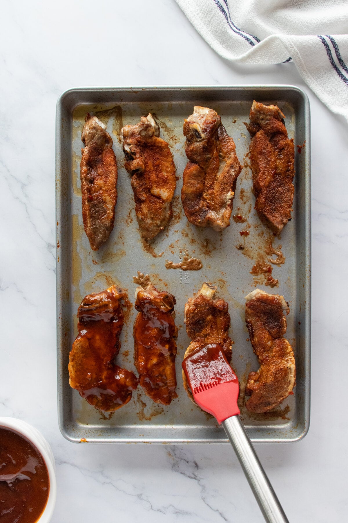 A baking sheet with eight pork rib pieces; four are coated with dry rub and four partially brushed with barbecue sauce using a red silicone brush. A small bowl of barbecue sauce and a white towel are nearby on a marble surface.