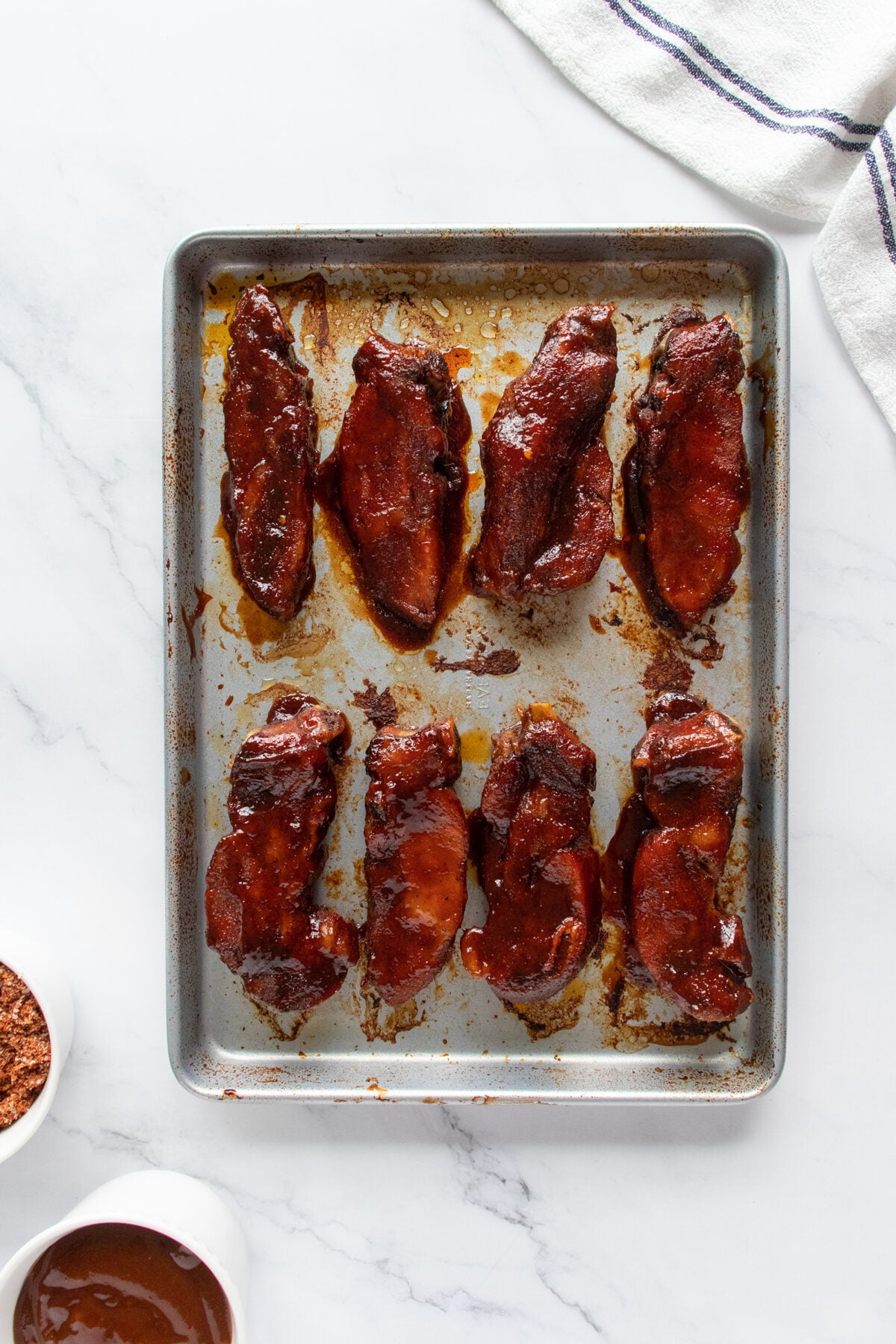 A baking sheet with eight glazed, cooked pork ribs arranged in two rows on a marble surface. A striped kitchen towel and small bowls with sauce and seasoning are nearby.